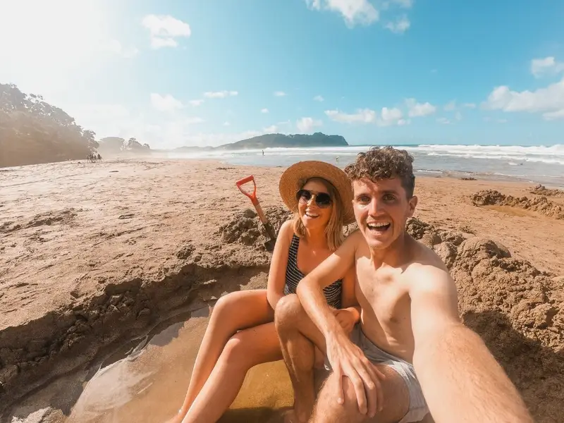 A couple smiles while sitting in a sand pool filled with warm water at Hot Water Beach on the North Island of New Zealand while on a spring small group tour.
