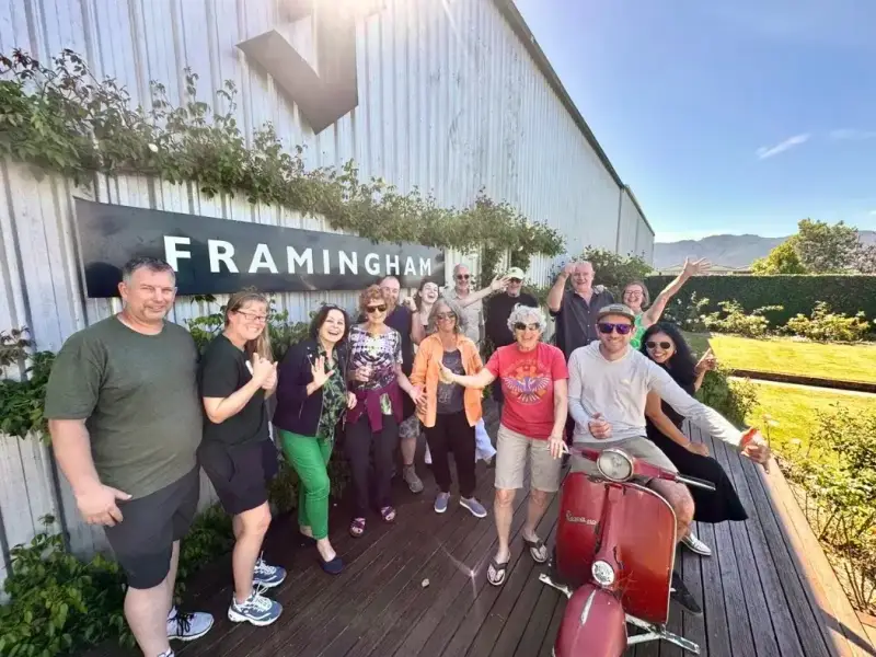 A group of Wild Kiwi travellers pose outside Framingham Winery in Marlborough, New Zealand, beside the winery sign on a sunny day in wine country. This photo shows the kind of relaxed and social vineyard experience visitors can enjoy in Marlborough, one of the best regions to try New Zealand Sauvignon Blanc, cellar door tastings, and scenic South Island food and wine travel.