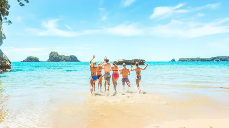 Group of young travellers running into clear turquoise water on a sunny New Zealand beach, capturing the excitement and freedom of summer adventures with Wild Kiwi Tours.