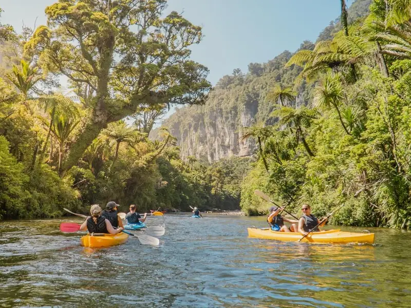 Group kayaking along a peaceful river surrounded by lush native forest and cliffs, showing why autumn in New Zealand is ideal for outdoor adventure days with comfortable temperatures and steady conditions.