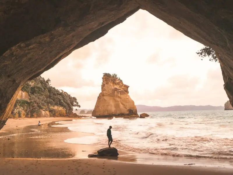 A fiery sunset at Cathedral Cove in the Coromandel Peninsula of the North Island of New Zealand in autumn, with the silhouette of a backpacker standing in the cave.