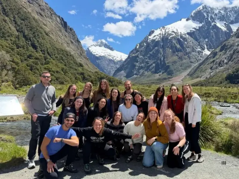 A group of travellers explore New Zealand in Autumn and pose together, smiling and happy, by a sun-soaked valley with mountains in the distance.