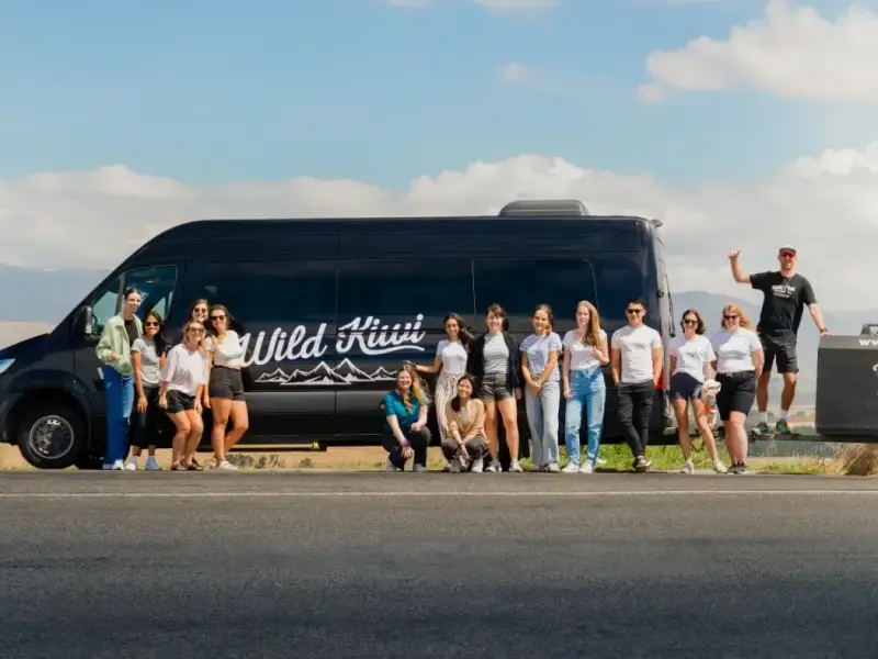 Wild Kiwi small-group travellers posing beside a Wild Kiwi tour van on a New Zealand road trip, showing the social, youthful vibe of guided group travel across Aotearoa.