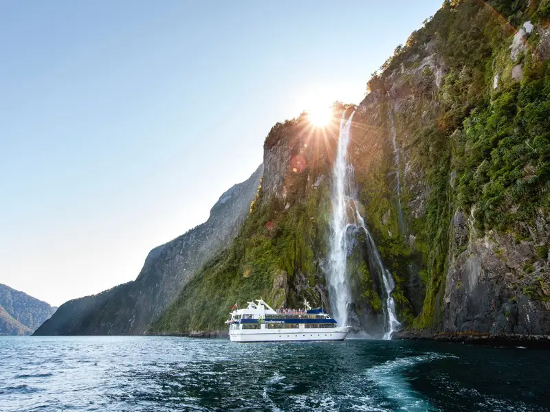 Photo of a luxury tour boat in Milford Sound in New Zealand under waterfalls with the sun peaking through.