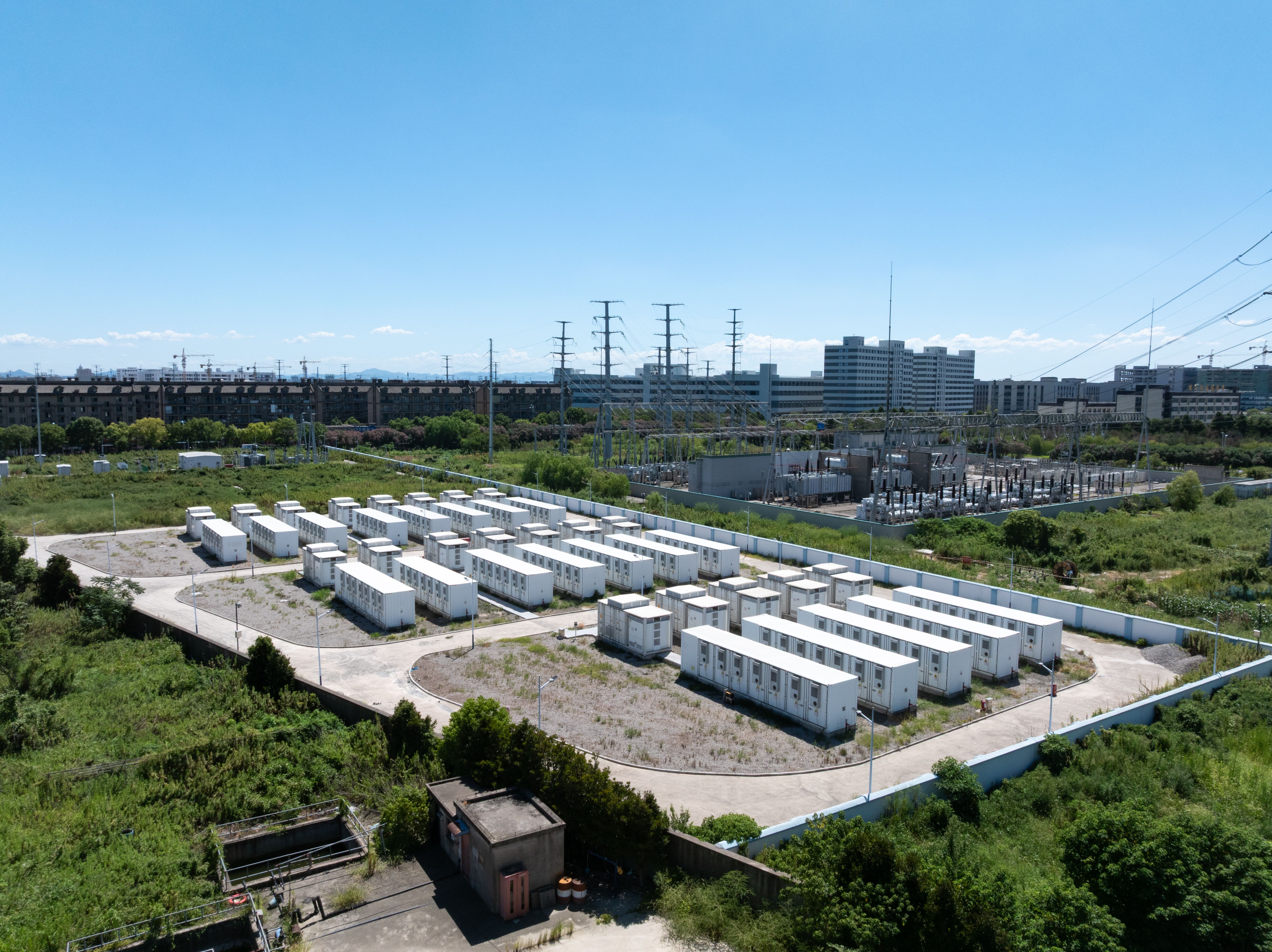 Aerial view of a grid-scale battery energy storage installation: rows of white battery container units beside an electrical substation under a clear blue sky