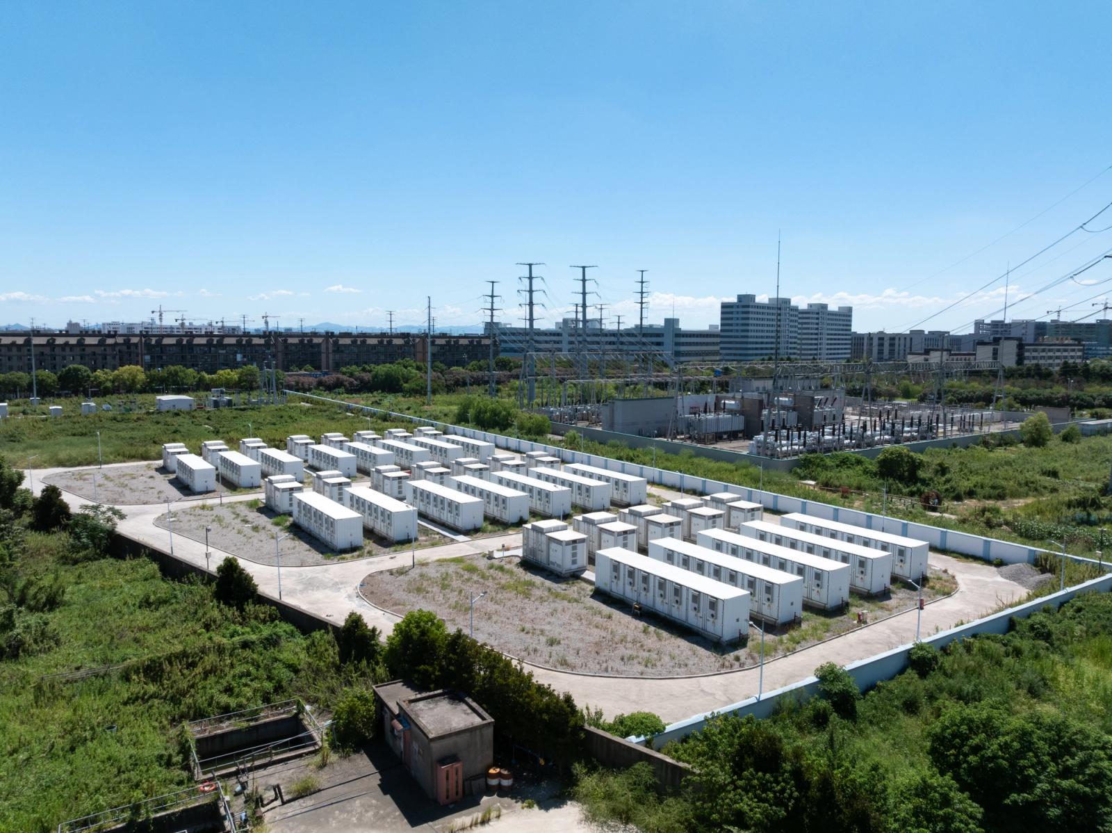 Aerial view of a grid-scale battery energy storage installation: rows of white battery container units beside an electrical substation under a clear blue sky