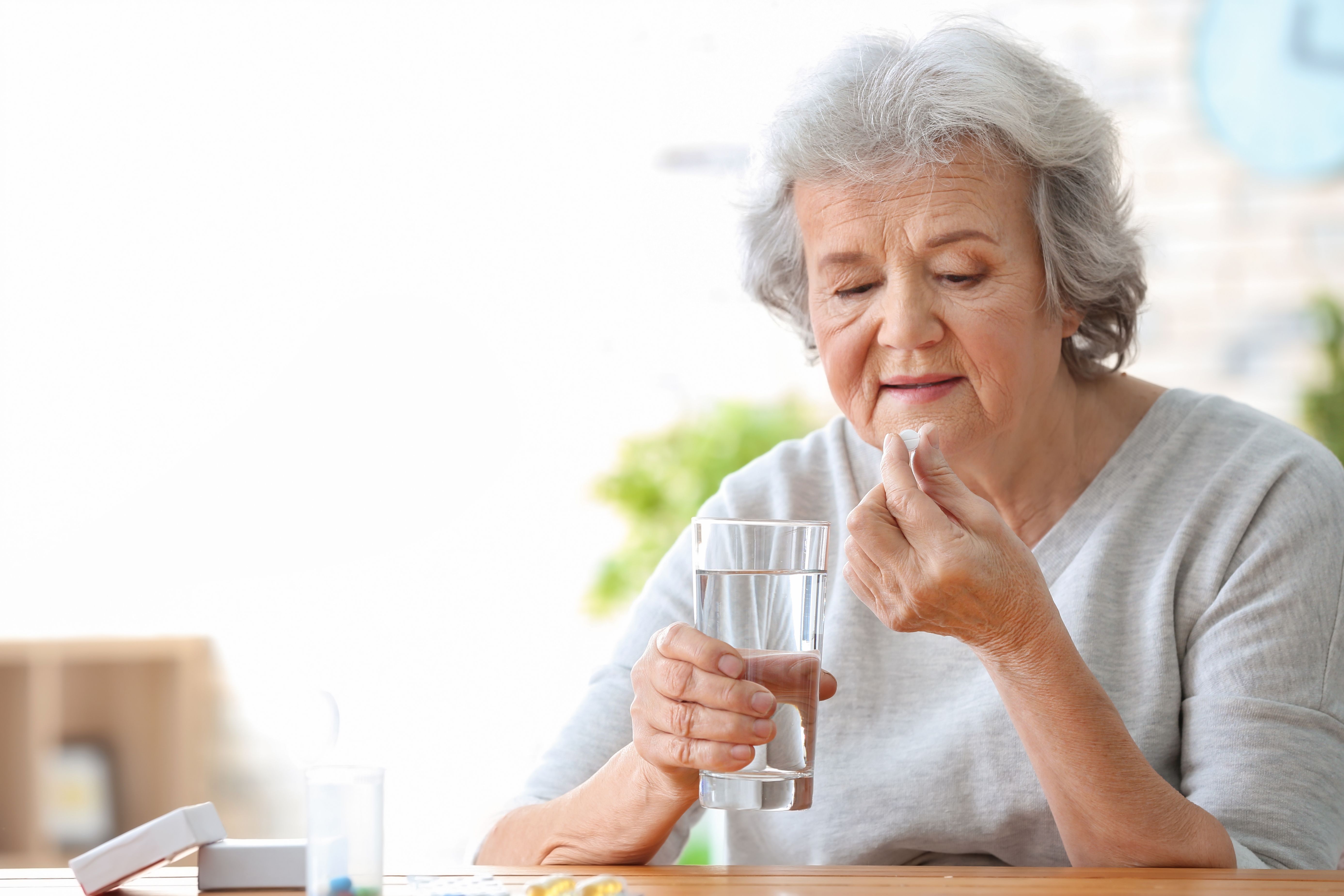 A woman in her seventies takes a tablet with a glass of water at a kitchen table.