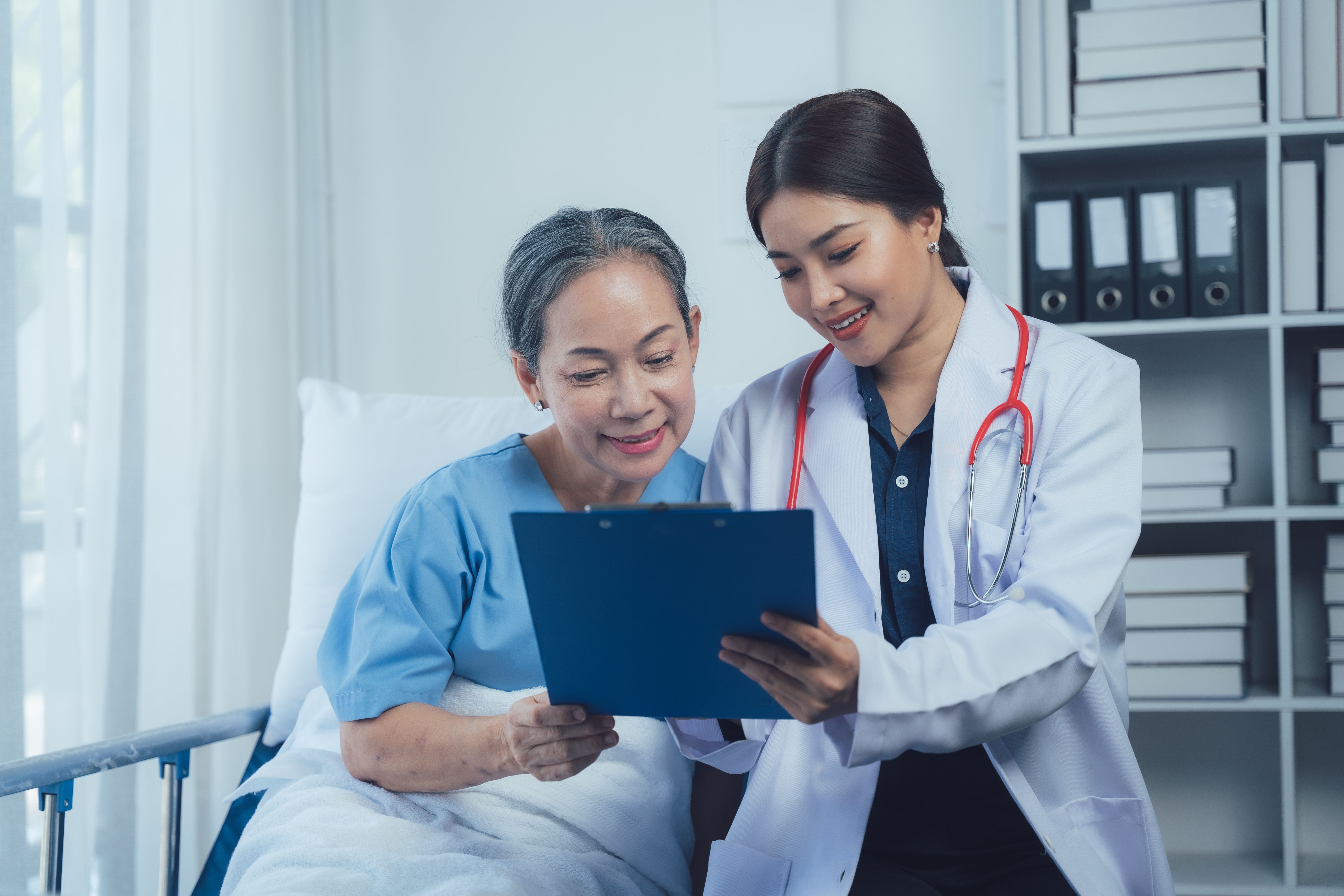 A doctor sits beside a smiling older woman in a hospital bed and shows her results on a tablet; a representative image of cancer care, not a specific patient