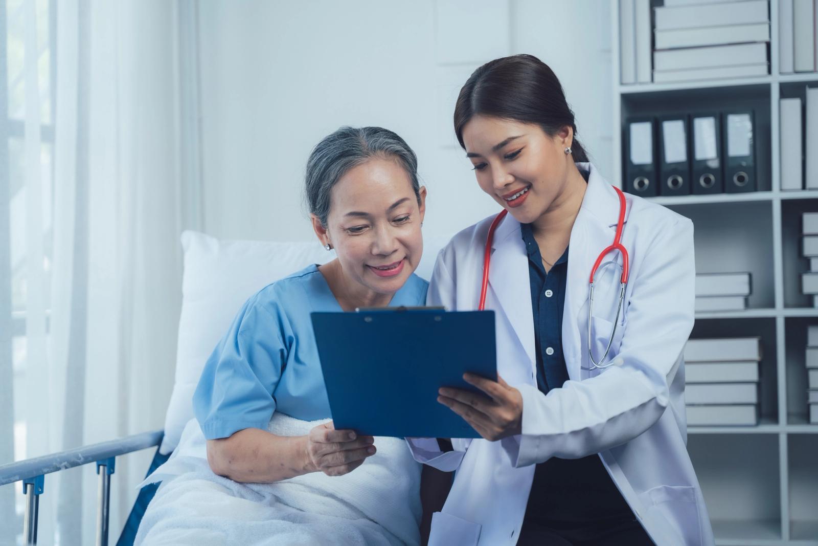 A doctor sits beside a smiling older woman in a hospital bed and shows her results on a tablet; a representative image of cancer care, not a specific patient