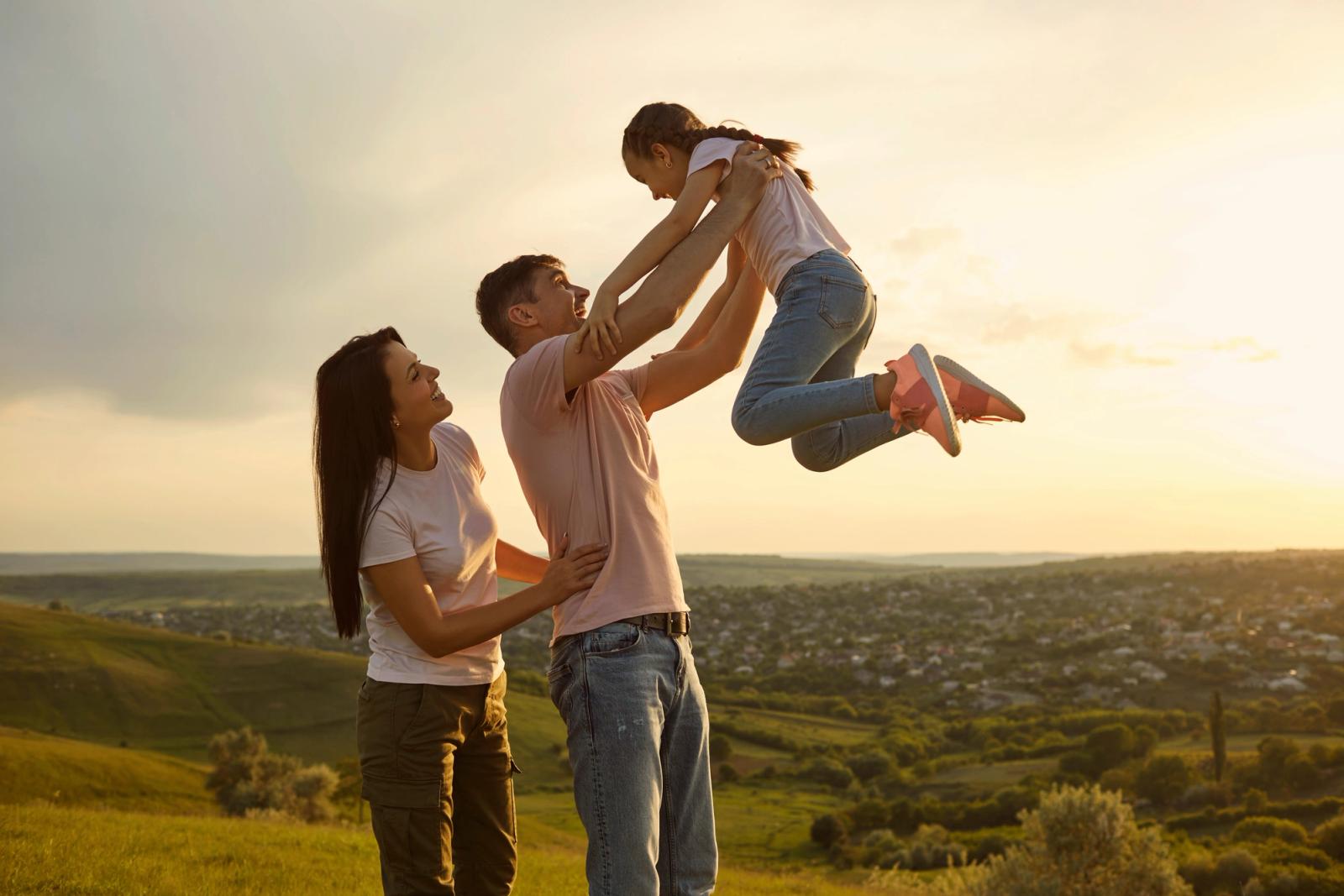 Parents lifting their laughing young daughter into the air outdoors at sunset