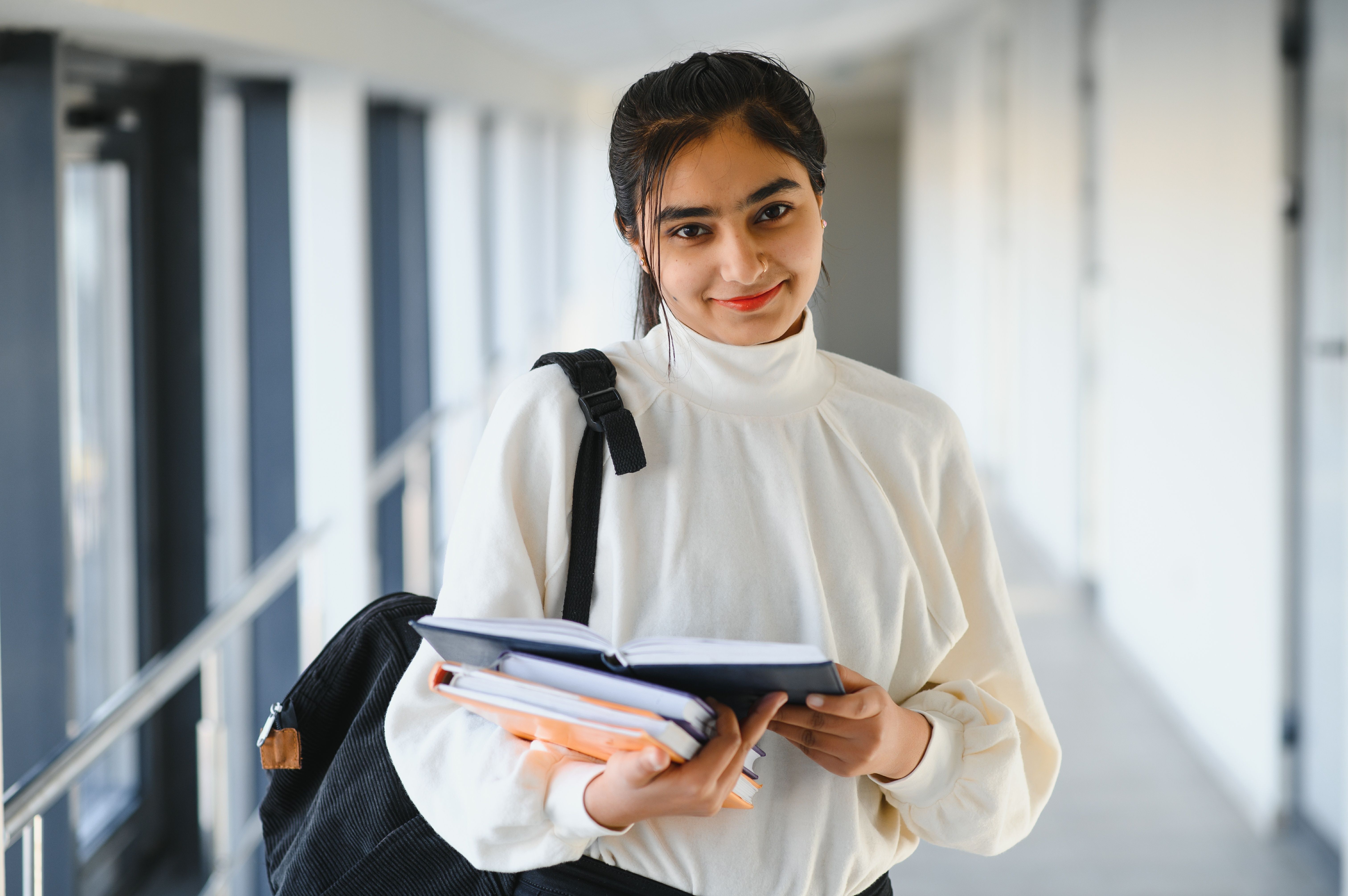 A young Indian woman student smiling and holding books in a bright college corridor