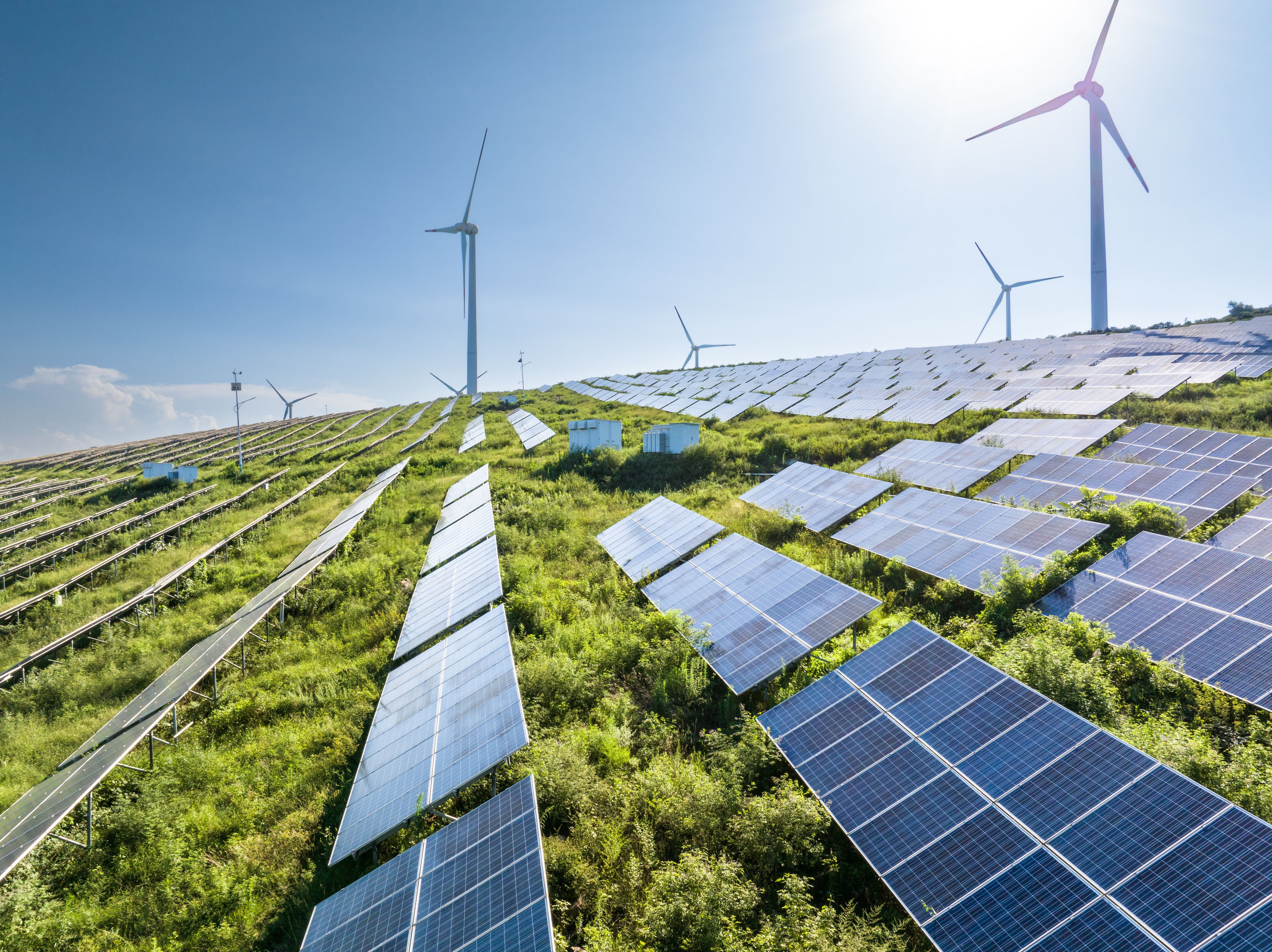 An aerial view of a hillside solar farm, rows of photovoltaic panels set among green vegetation with wind turbines along the ridge