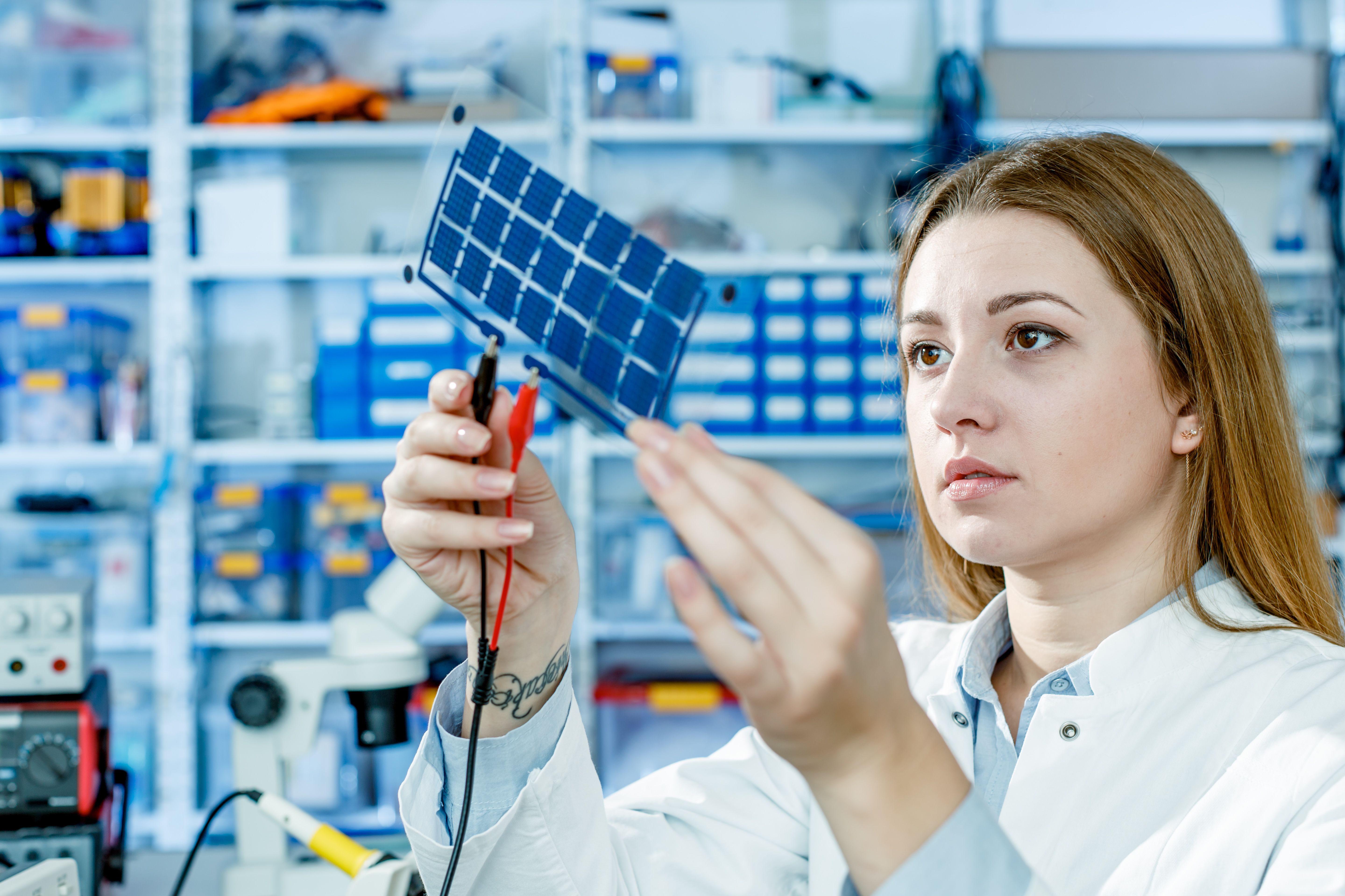 A researcher in a laboratory holds a thin, flexible solar cell fitted with test leads, examining it up close.