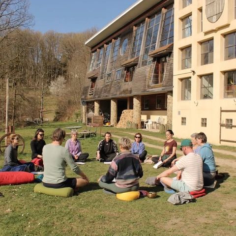 Deelnemers zitten in een cirkel op meditatiekussens in het gras voor een houten retreatlocatie