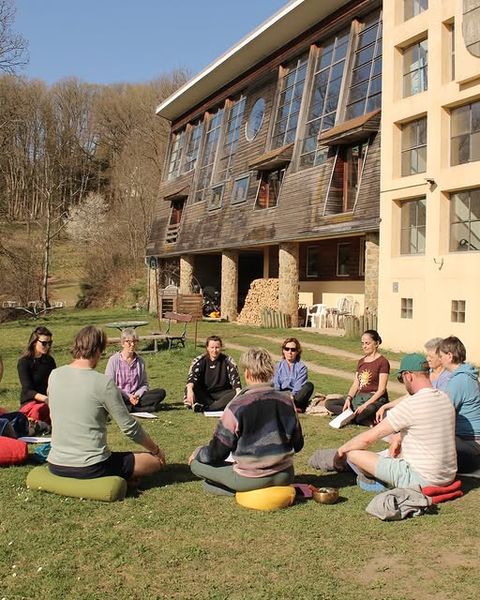 Deelnemers zitten in een cirkel op meditatiekussens in het gras voor een houten retreatlocatie