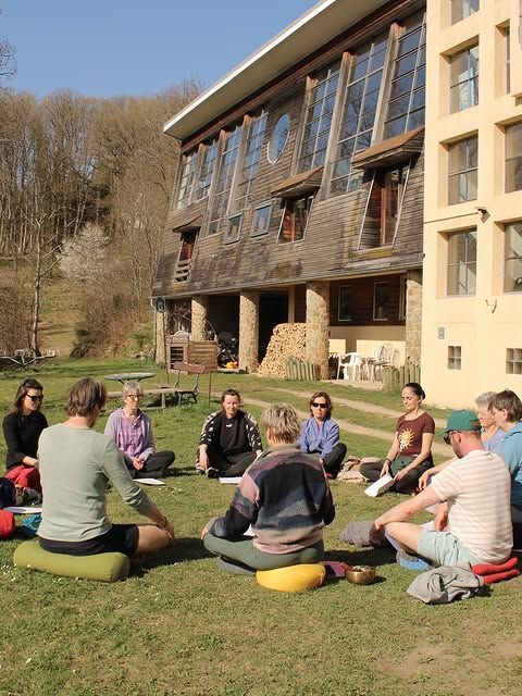 Deelnemers zitten in een cirkel op meditatiekussens in het gras voor een houten retreatlocatie
