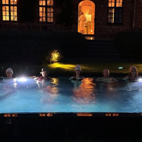 Vijf mensen met wintermutsen tot aan hun schouders in een verlicht zwembad in de avond