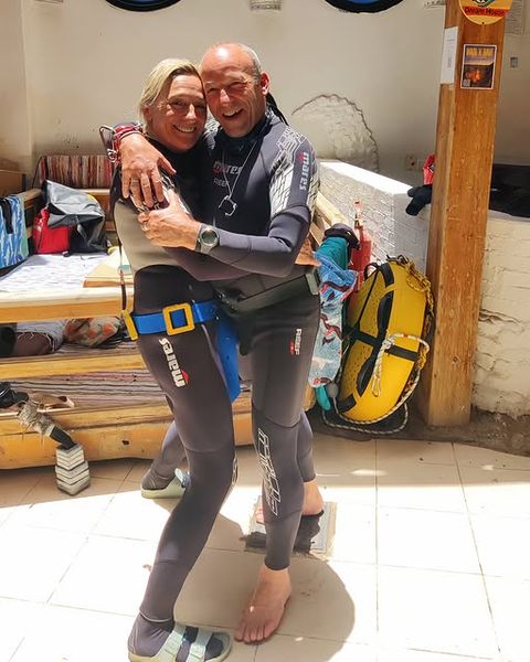 Rita en Philippe omhelzen elkaar glimlachend in duikpakken op een zonnig terras
