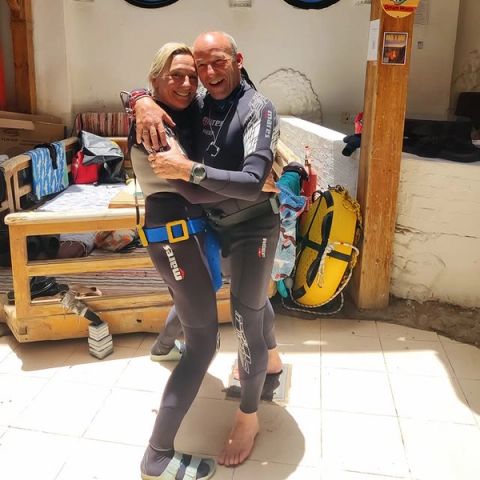 Rita en Philippe omhelzen elkaar glimlachend in duikpakken op een zonnig terras