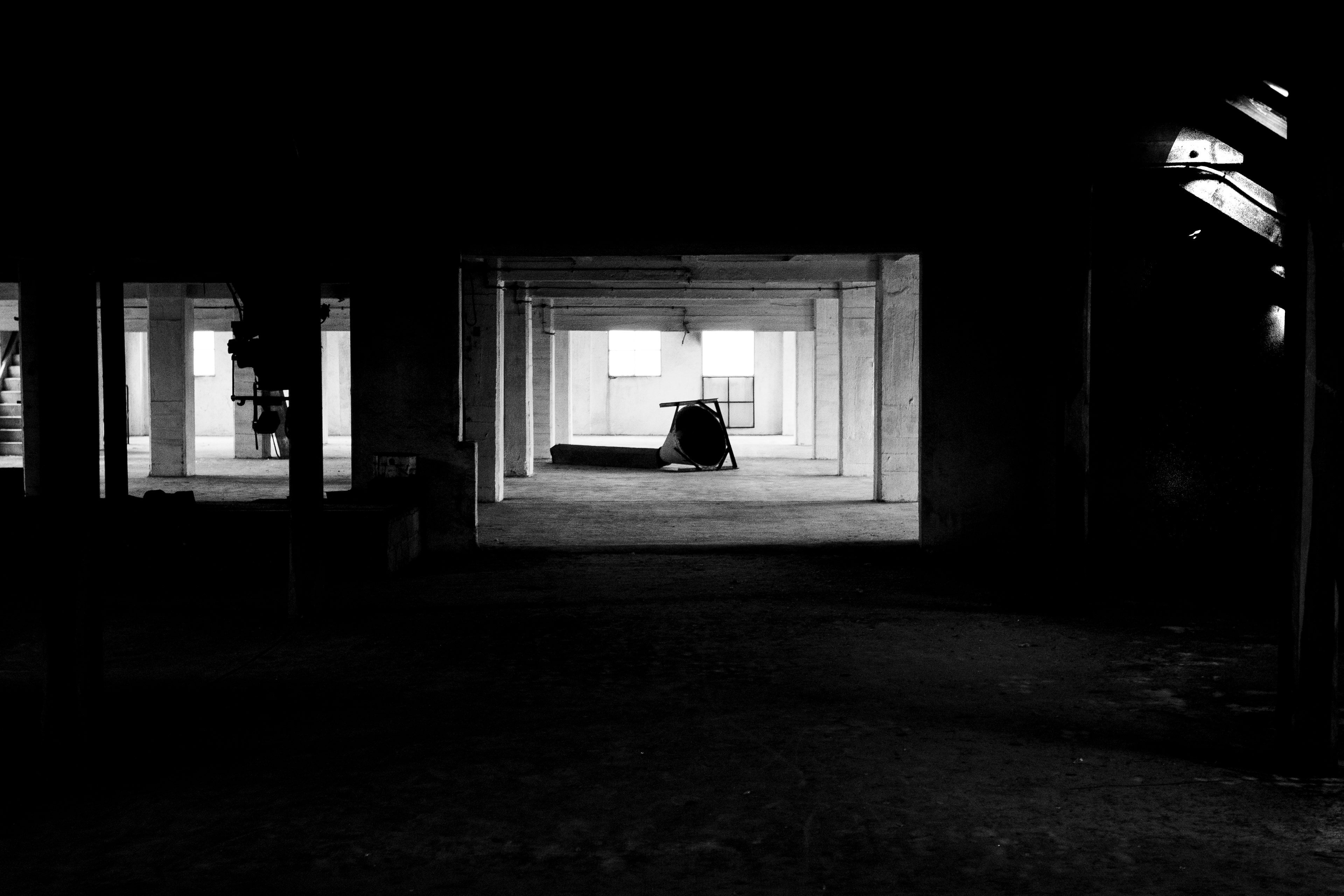 Black and white urbex photograph inside an old abandoned mill in the Loiret, France, with deep shadows, concrete pillars, empty industrial space and bright windows in the distance.