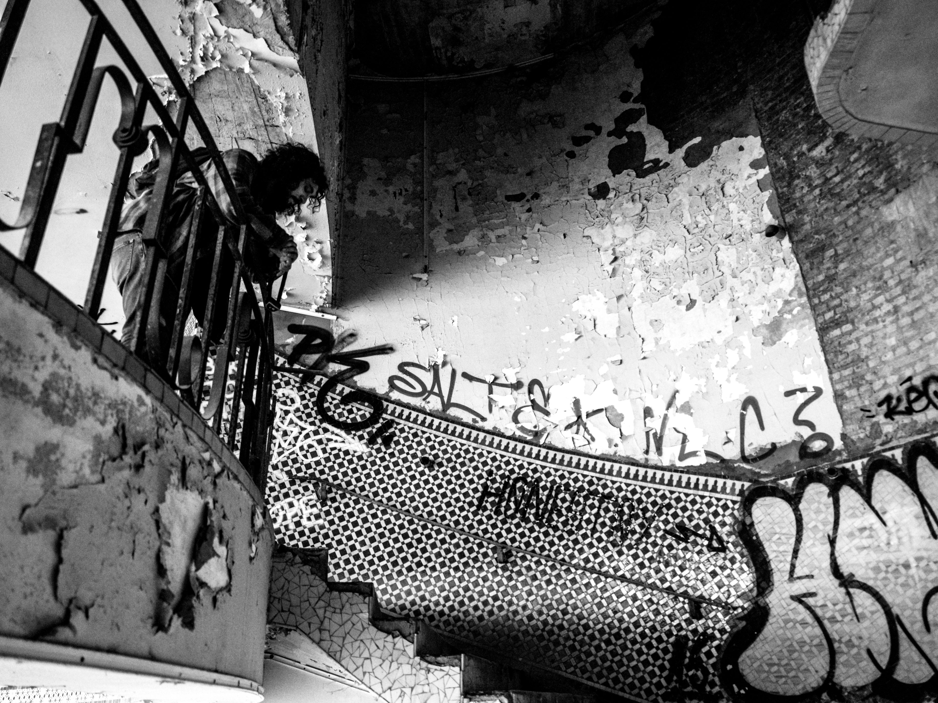 Black and white urbex photograph of a solitary figure near a damaged stairway inside an abandoned swimming pool in Charleroi, Belgium, with peeling walls, broken tiles, graffiti and strong contrast.