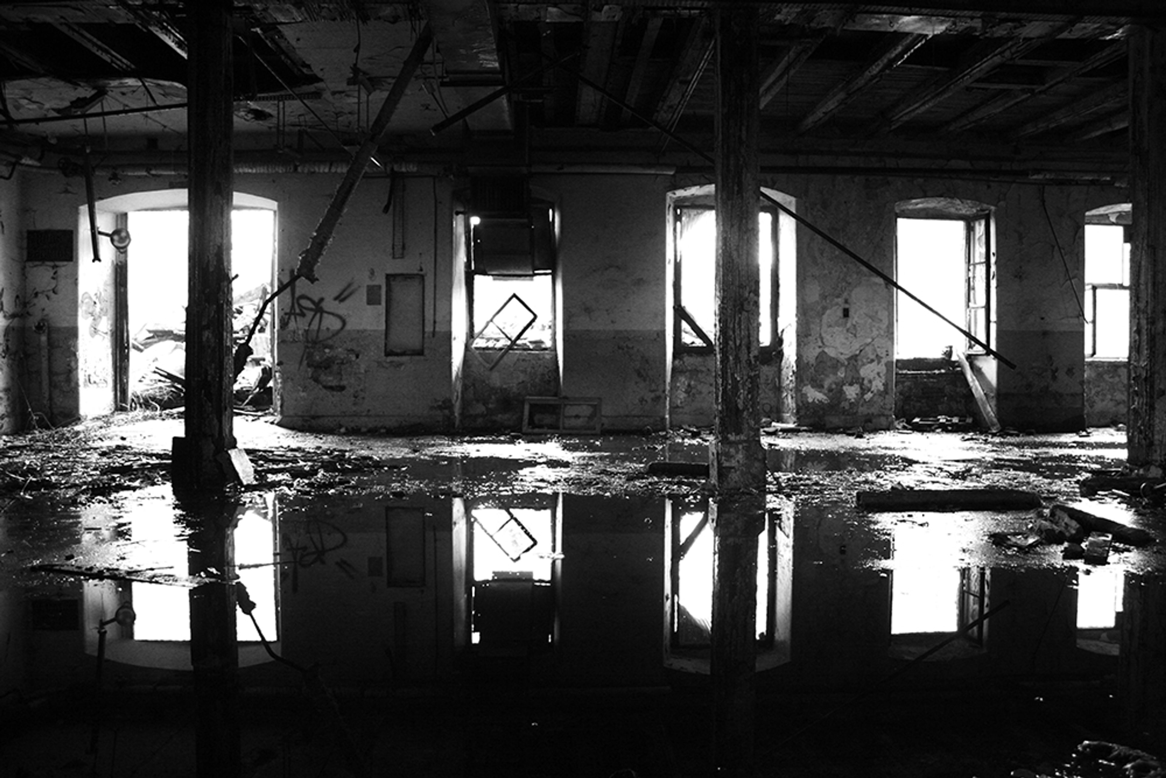 Black and white urbex photograph inside a fire-damaged factory in Mulhouse, France, showing standing water on the floor, broken windows, dark structural columns and a charred industrial interior.