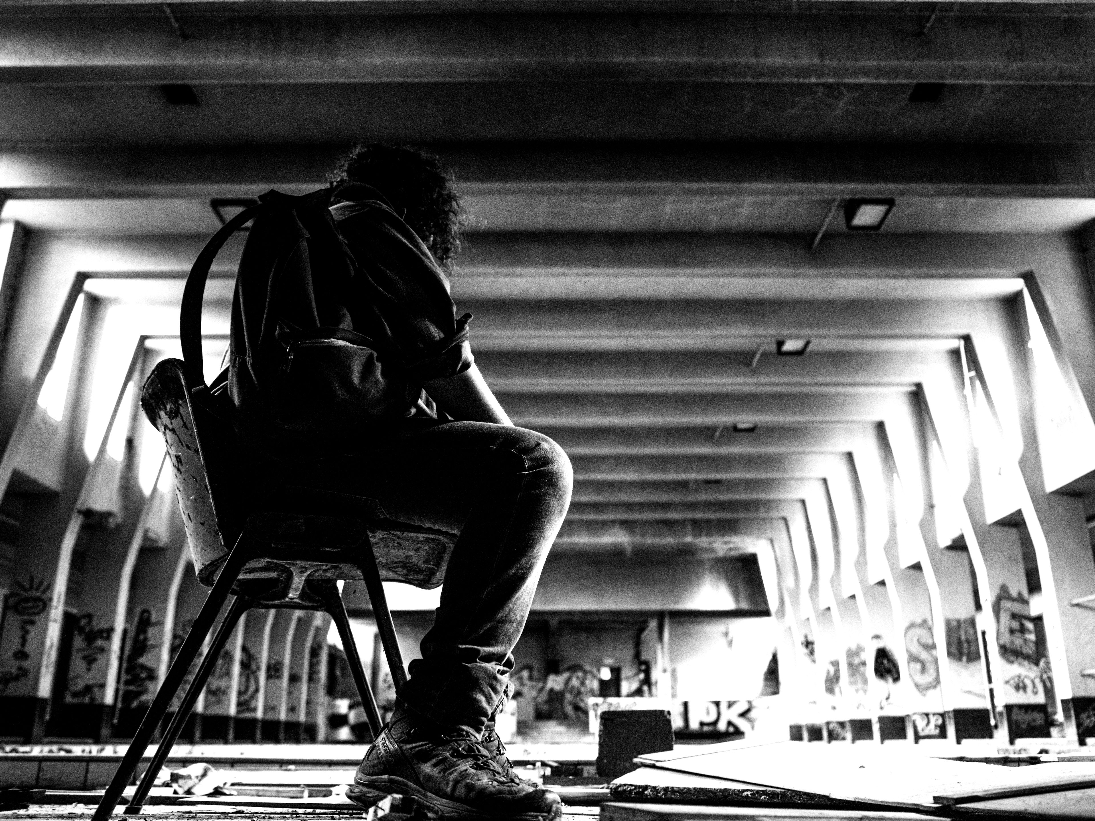 Black and white urbex photograph of a seated figure inside an abandoned swimming pool in Charleroi, Belgium, with graffiti-covered walls, empty tiled basins, strong backlight and decaying interior architecture.