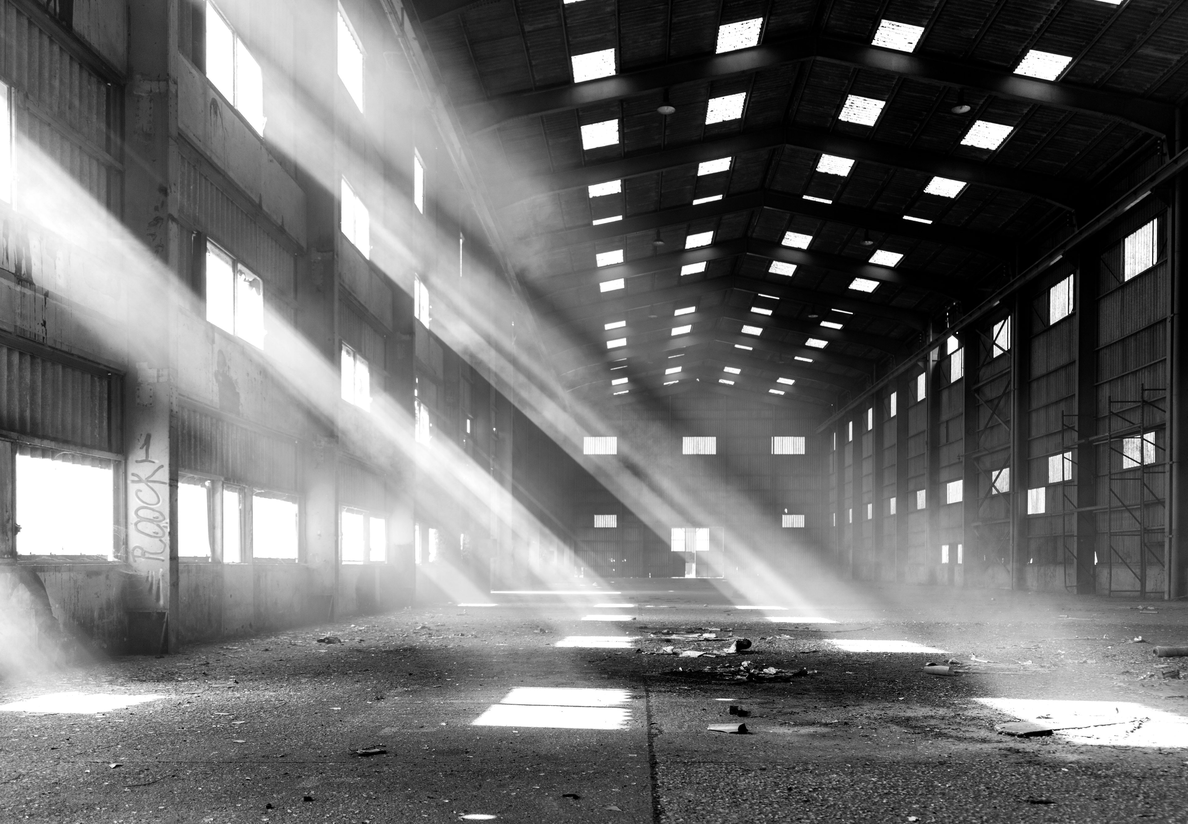 Black and white urbex photograph inside an abandoned factory in Clamecy, France, with dramatic beams of sunlight entering through high windows, an empty industrial hall and a large metal roof structure.