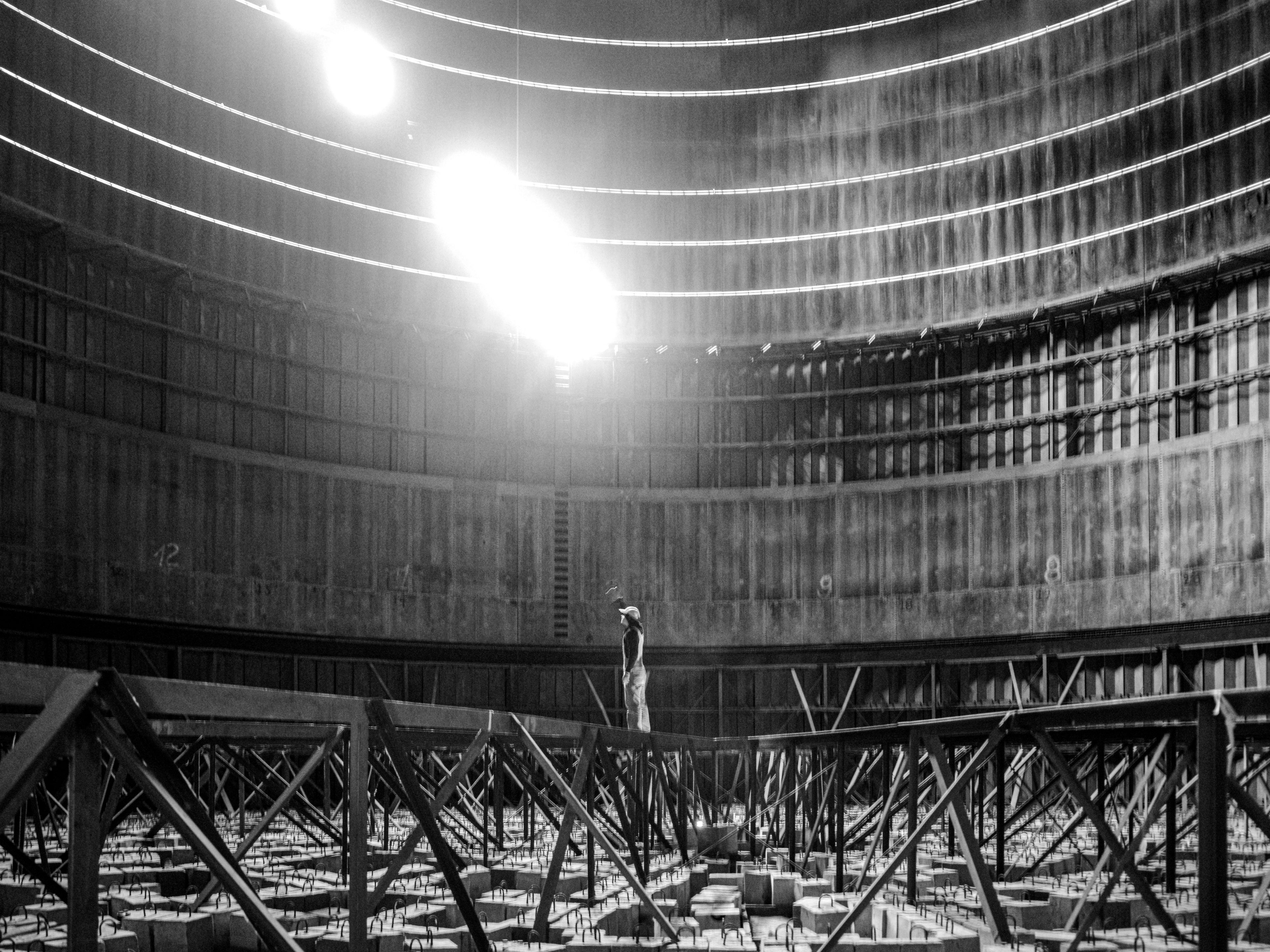 Black and white urbex photograph inside an abandoned reservoir in Charleroi, Belgium, near a thermal power plant, with circular concrete walls, metal structures, bright overhead light and a lone figure standing inside.