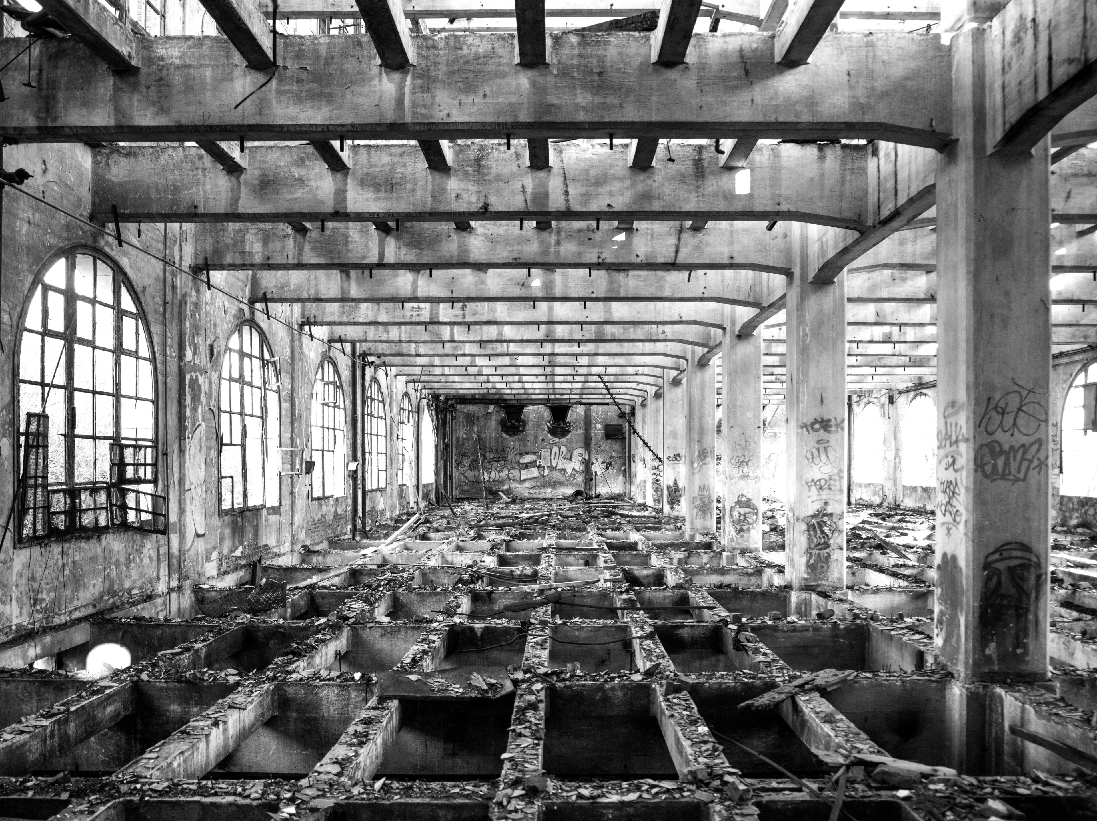 Black and white urbex photograph of the empty concrete structure inside the former mill in Marquette-lez-Lille, with exposed beams, arched windows, graffiti and an abandoned industrial floor.