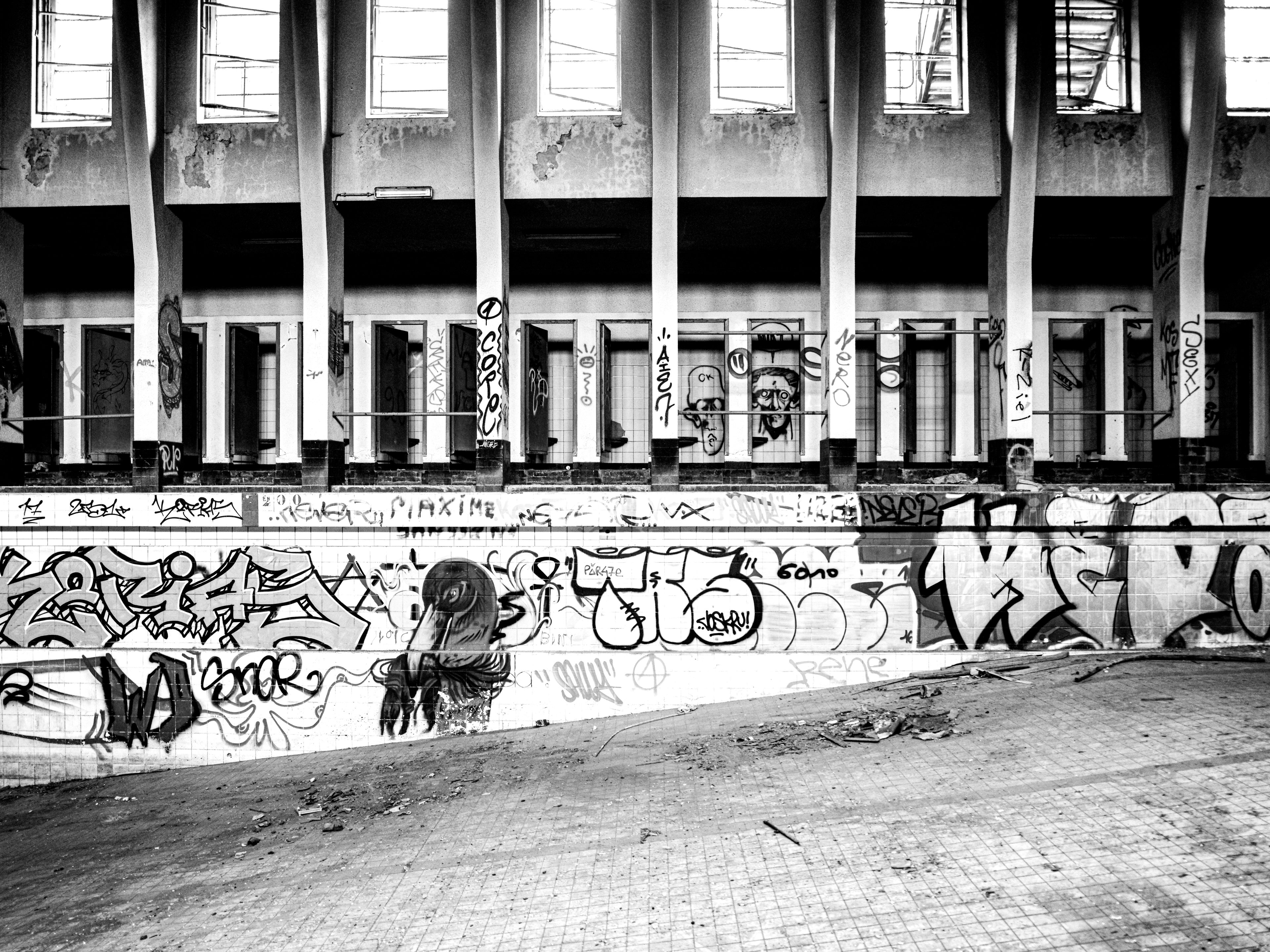 Black and white urbex photograph of graffiti-covered walls inside an abandoned swimming pool in Charleroi, Belgium, with empty changing areas, tiled surfaces, tall columns and decaying interior architecture.