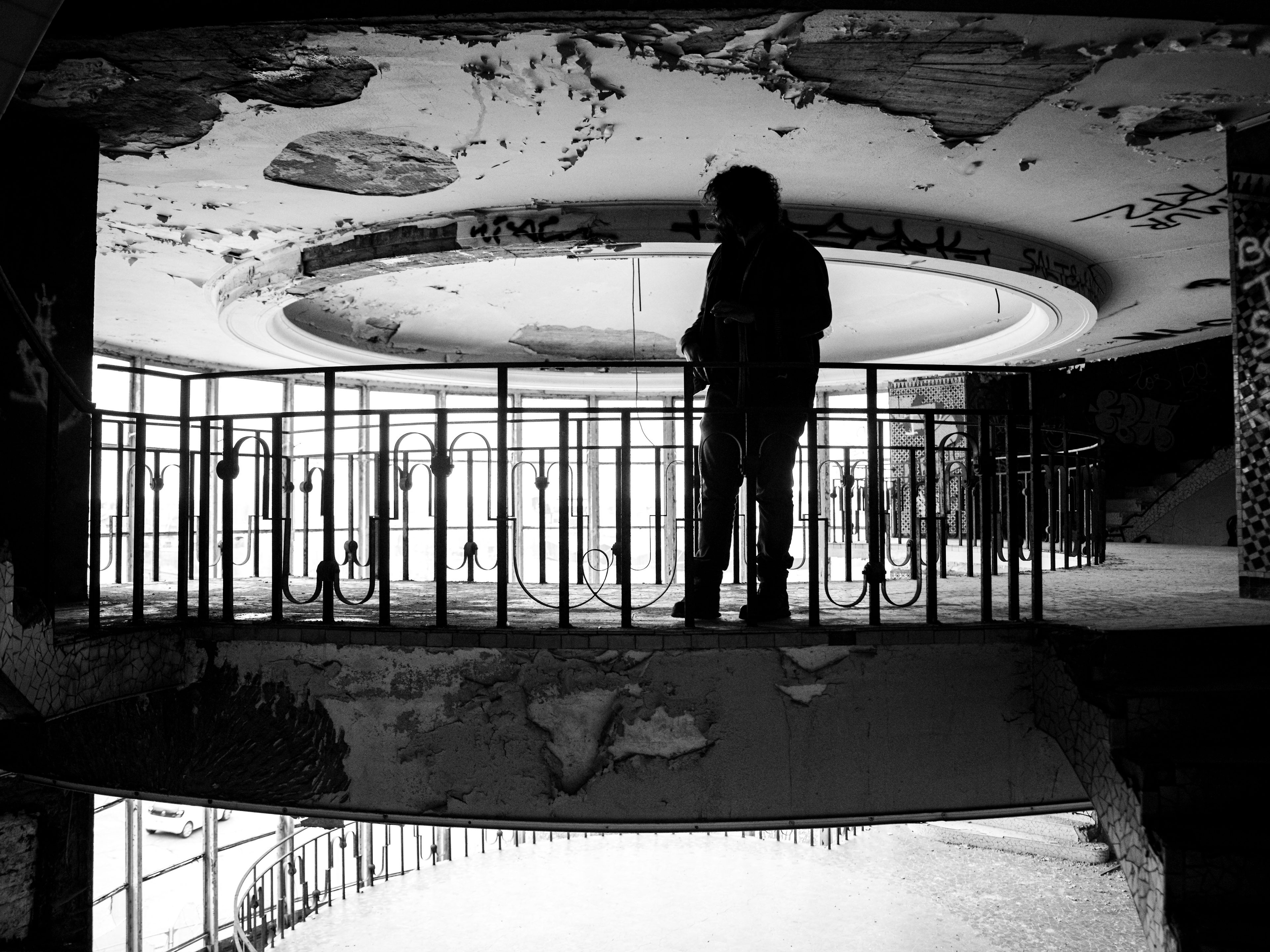 Black and white urbex photograph of a solitary silhouette inside an abandoned swimming pool in Charleroi, Belgium, with a decaying circular ceiling, metal railings, broken walls and strong backlight.