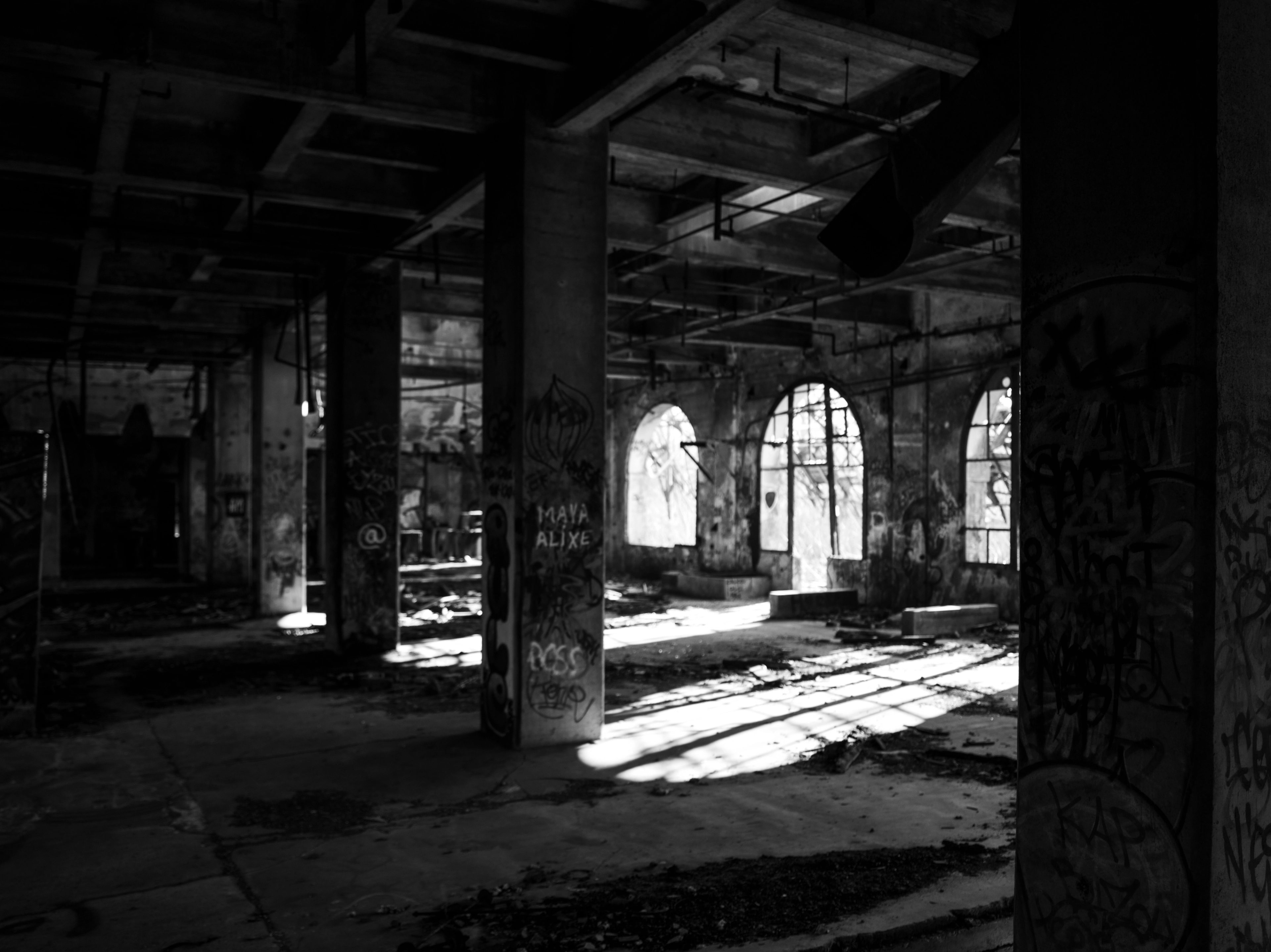 Black and white urbex photograph of the abandoned former Moulin de Paris interior in Marquette-lez-Lille, with arched windows, graffiti-covered concrete columns and sunlight on the floor.