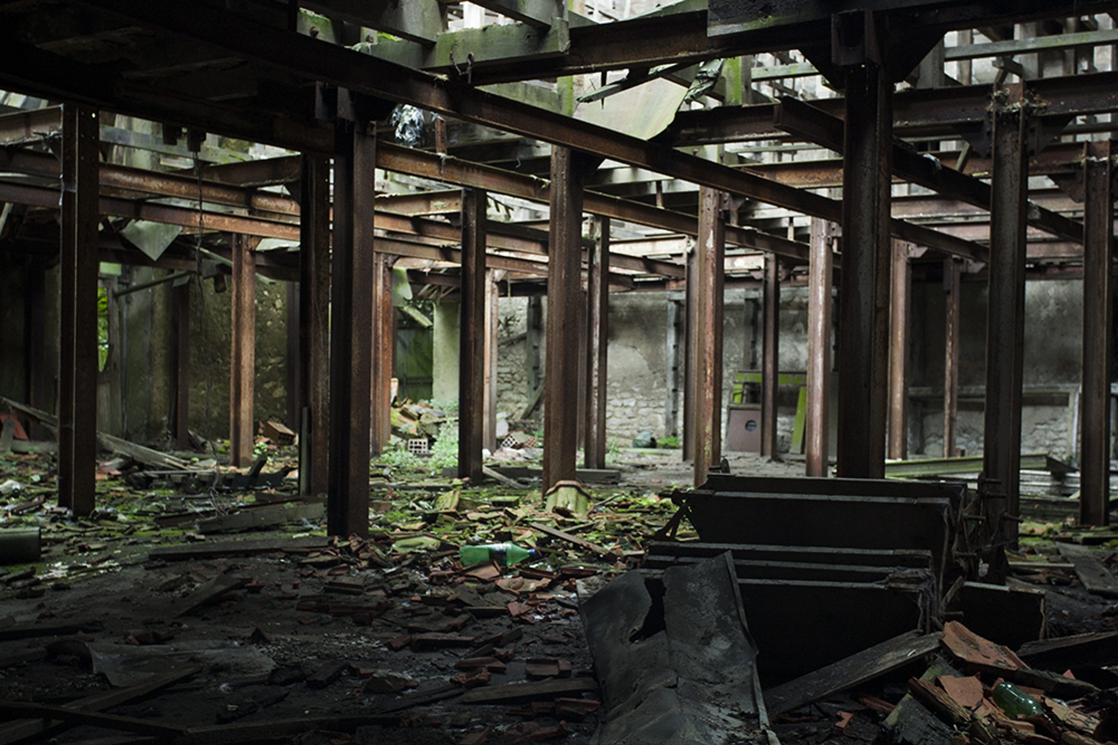 Color urbex photograph inside an abandoned factory in Vierzon, France, showing rusted metal beams, scattered debris, broken materials, vegetation and decaying industrial architecture.