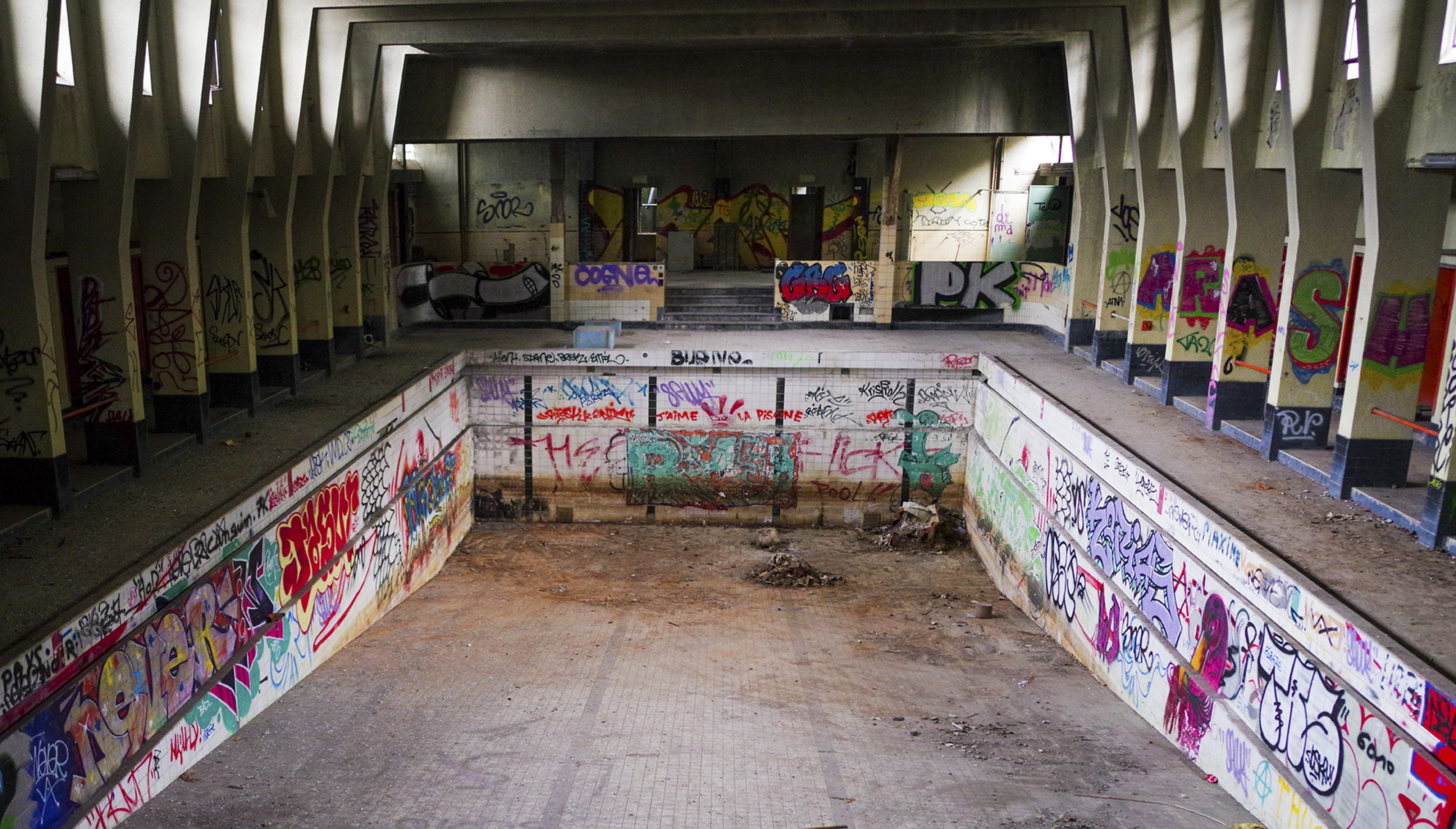 Color urbex photograph of an abandoned swimming pool in Charleroi, Belgium, with empty tiled basins, graffiti-covered walls, concrete columns and decaying interior architecture.