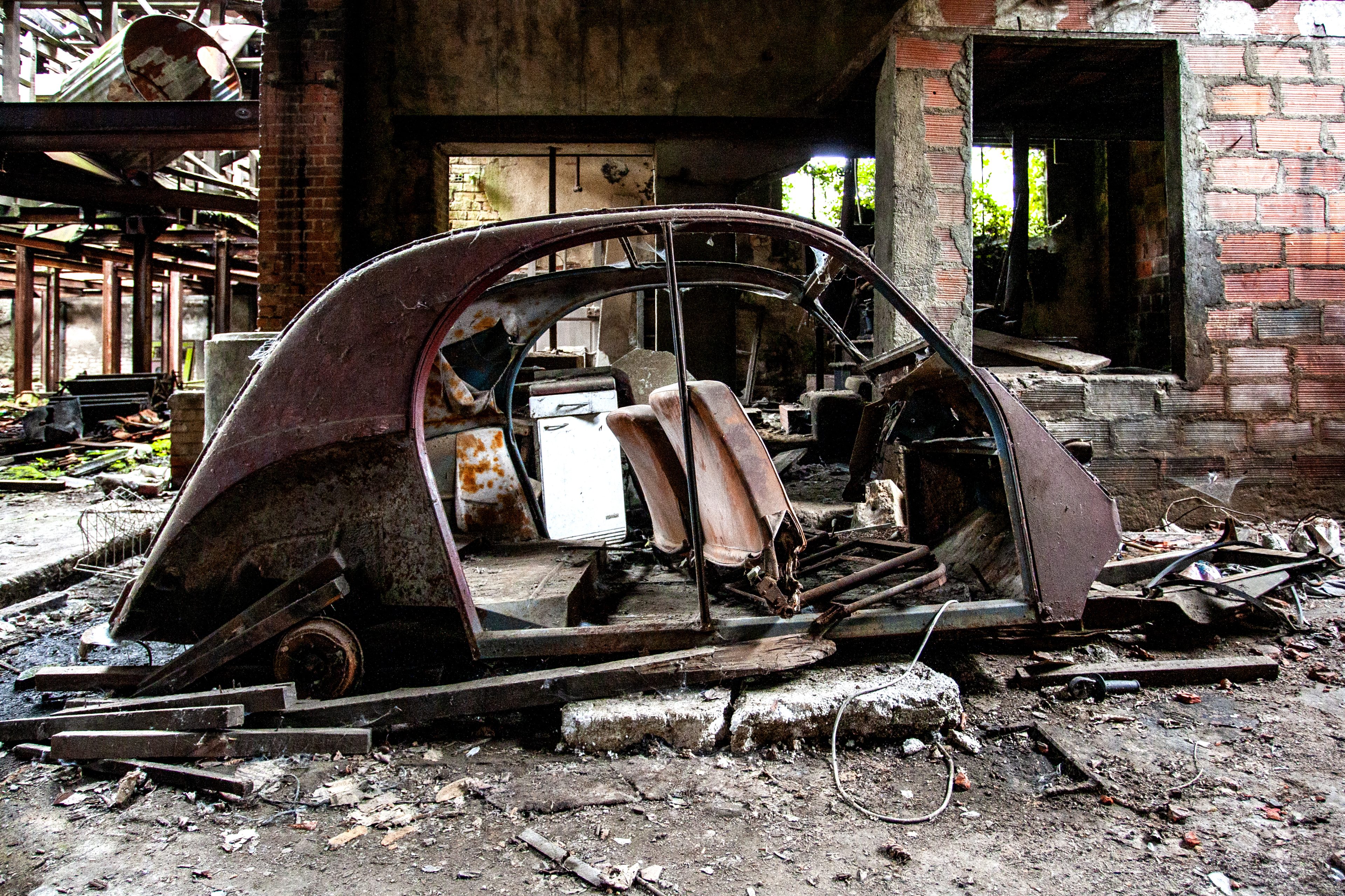 Color urbex photograph inside an abandoned factory in Vierzon, France, showing the shell of an old car surrounded by rusted metal, broken bricks, debris and decaying industrial walls.