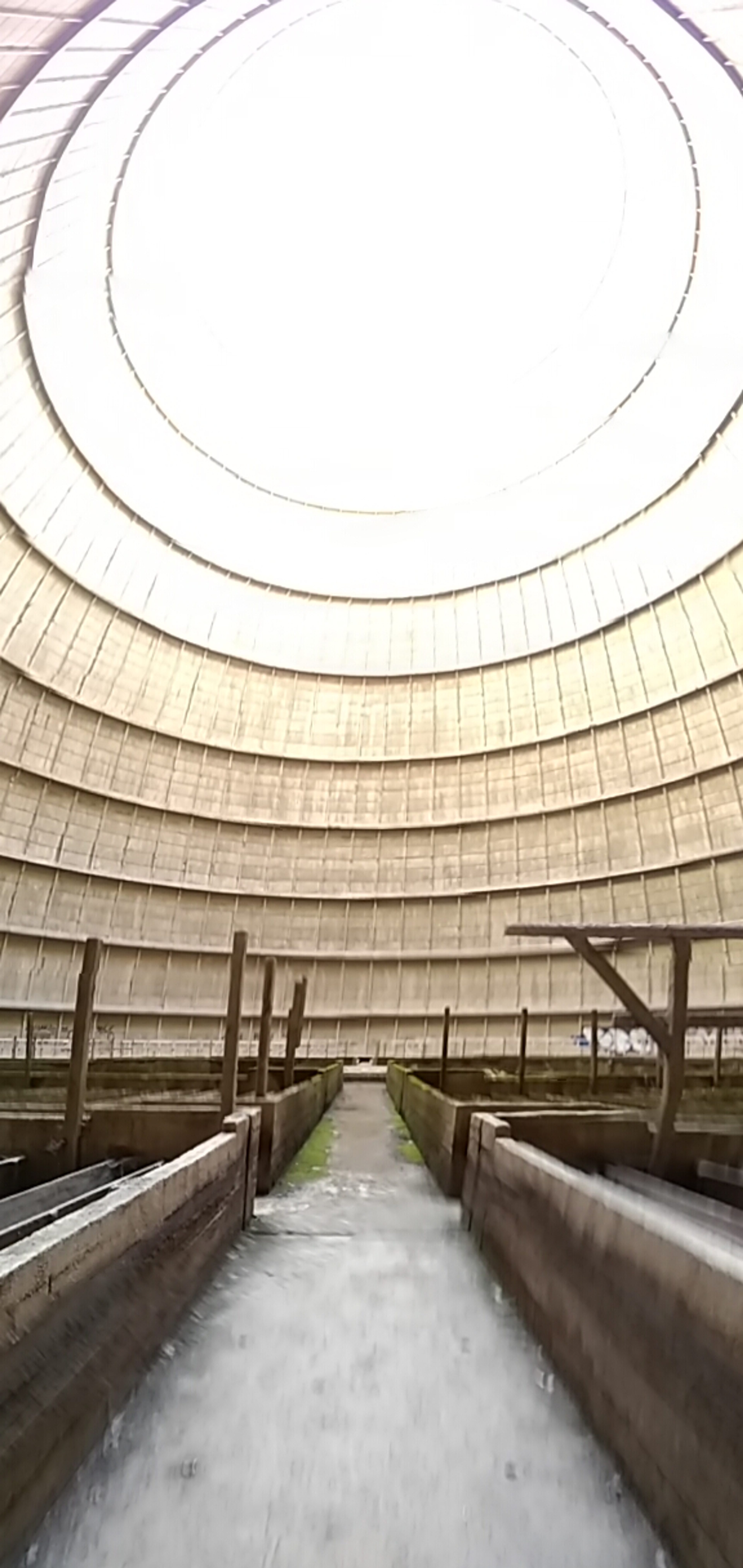 Vertical urbex photograph inside an abandoned cooling tower in Charleroi, Belgium, showing a circular concrete wall, bright open sky, industrial basins and a central pathway.