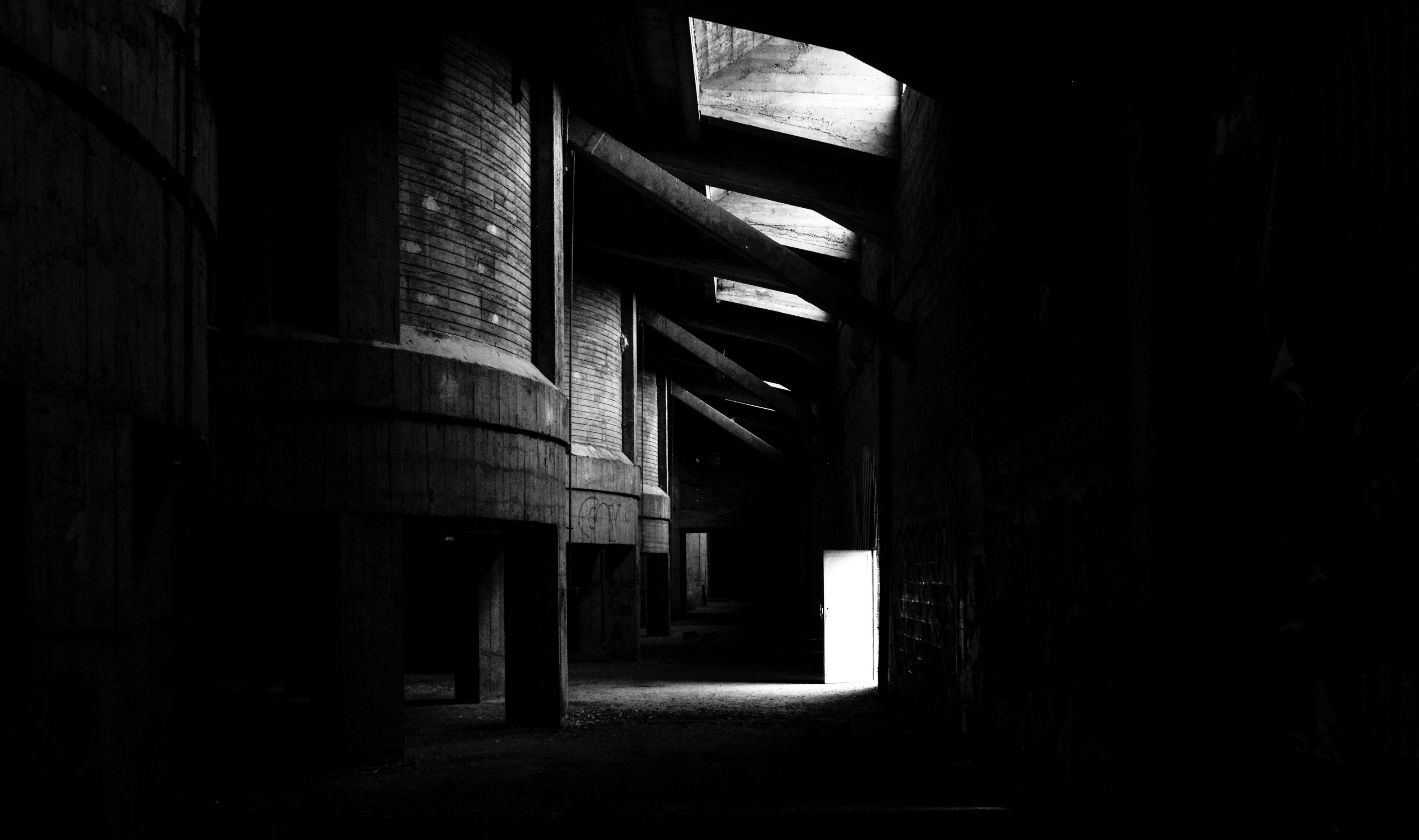 Black and white urbex photograph inside an abandoned silo in Tournai, Belgium, showing a dark industrial corridor, concrete structures, overhead openings and a bright doorway in the distance.