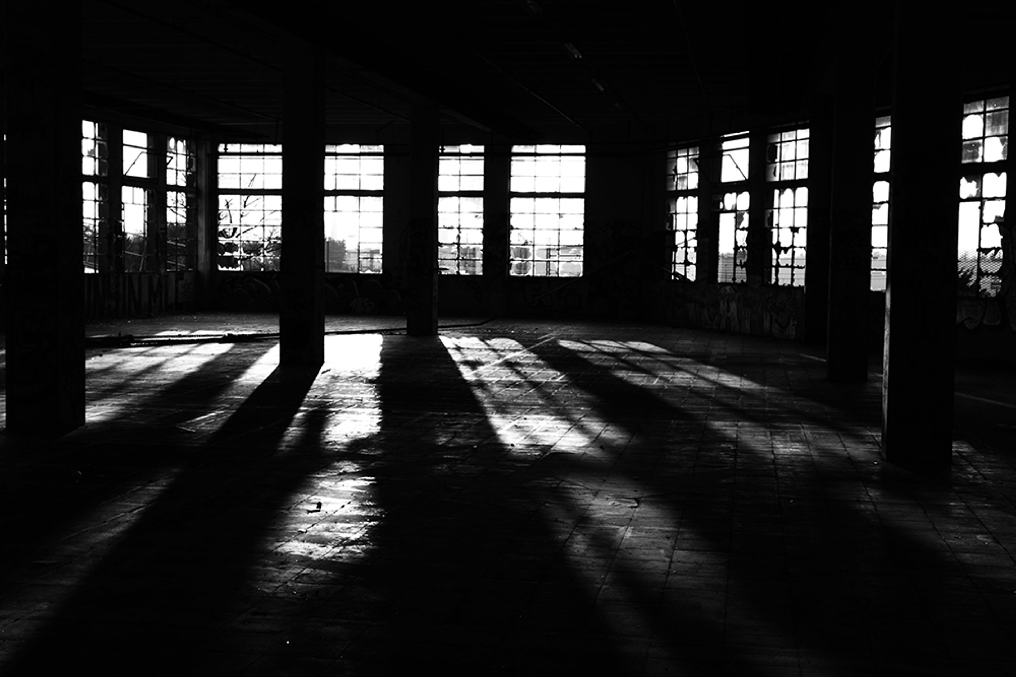 A black and white urbex photograph captured inside an abandoned factory in Orléans, France, where broken windows, concrete pillars and long shadows create a dark and silent industrial atmosphere.