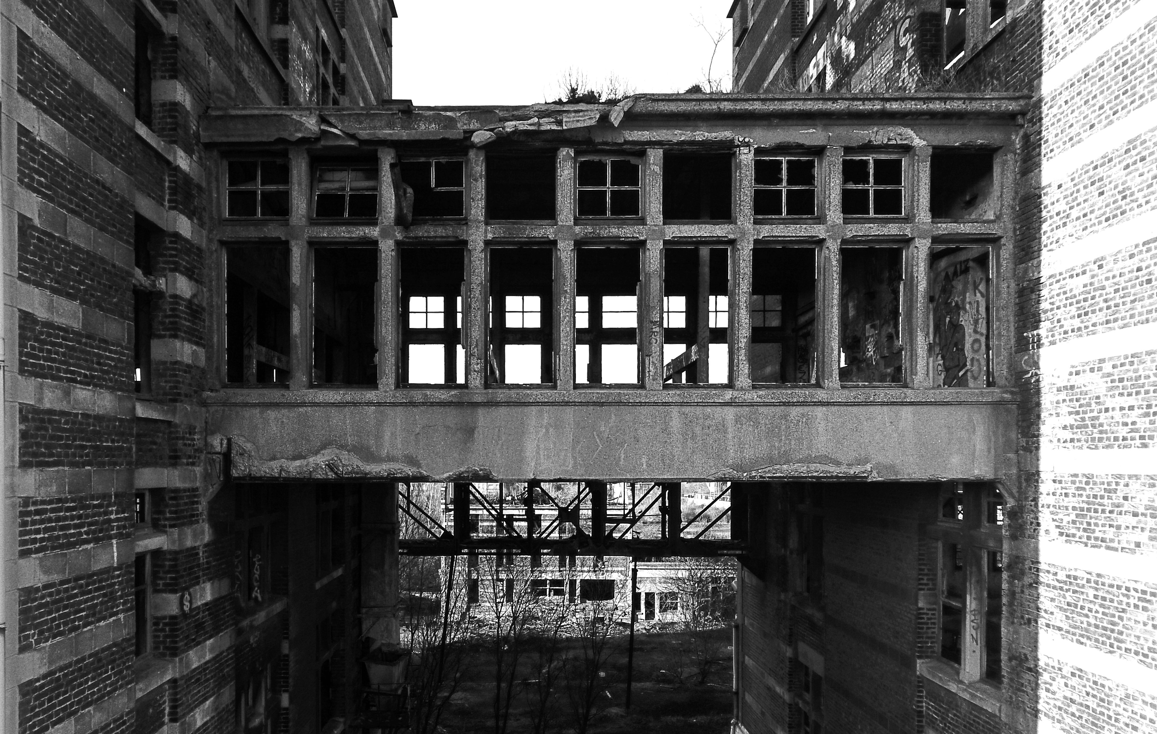 Black and white urbex photograph of an abandoned industrial passage in Marquette-lez-Lille, with brick walls, empty windows, concrete beams and a suspended structure between old buildings.