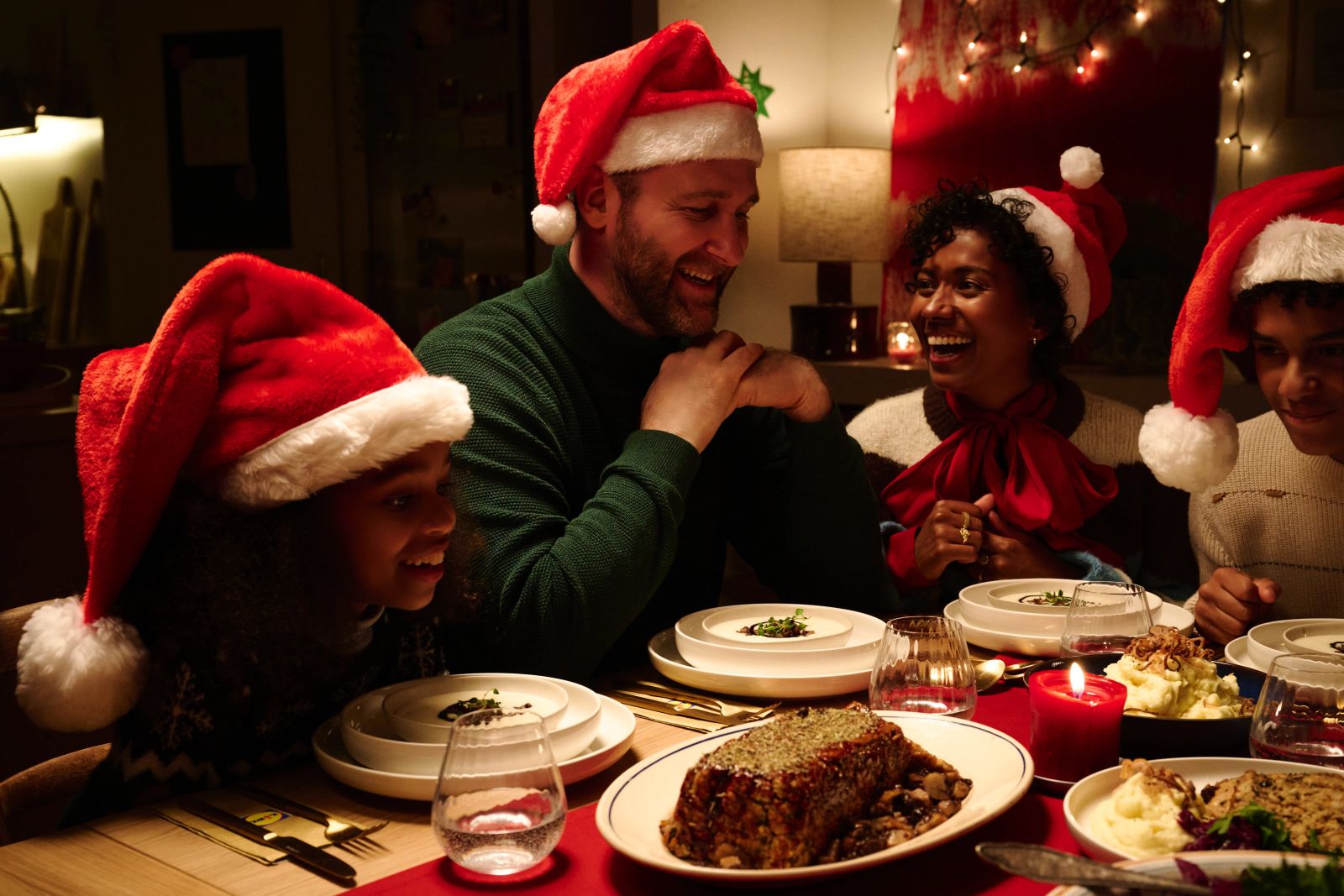 Eine Gruppe von Personen mit roten Weihnachtsmützen sitzt lachend und essend an einer festlich gedeckten Tafel