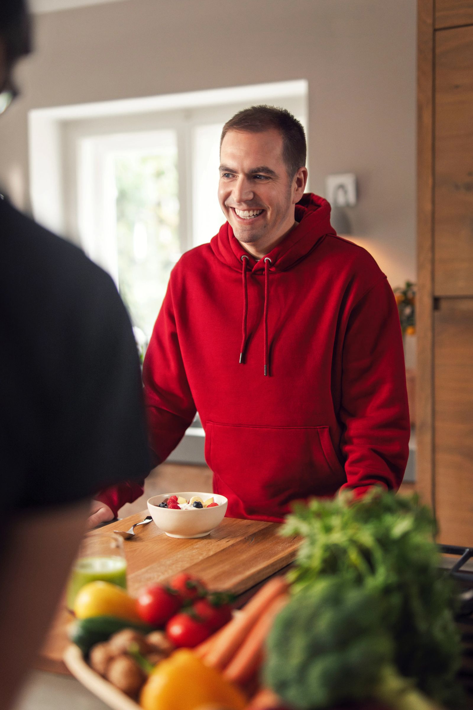 Philipp Lahm steht lächelnd in einer modernen Holzküche und bereitet ein gesundes Müsli zu. Auf der Arbeitsplatte liegen frisches Obst und Gemüse wie Bananen und Brokkoli bereit. Fotografiert von Karl Scholten.