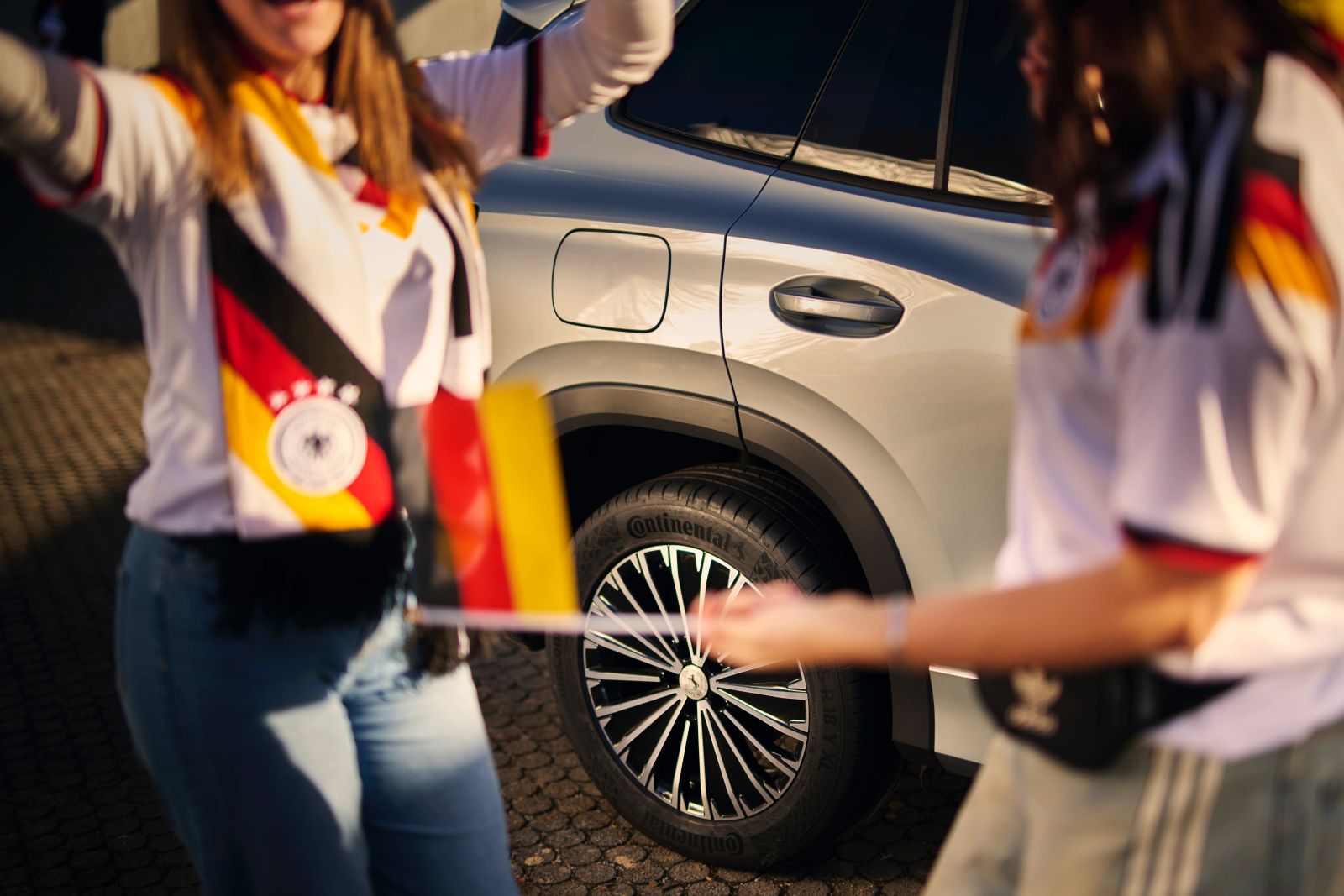 Zwei ausgelassene Fans in Deutschland-Trikots und mit Fanutensilien jubeln vor einem silbernen Continental-Fahrzeug; Werbemotiv für die Continental WM 2026 Kampagne, fotografiert von Karl Scholten