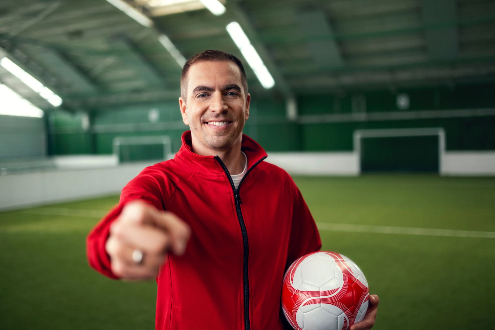 Philipp Lahm steht lächelnd in einer modernen Fussballhalle und zeigt mit einer Hand in die Kamera, in der anderen Hand hält er einen Ball. Fotografiert von Karl Scholten.