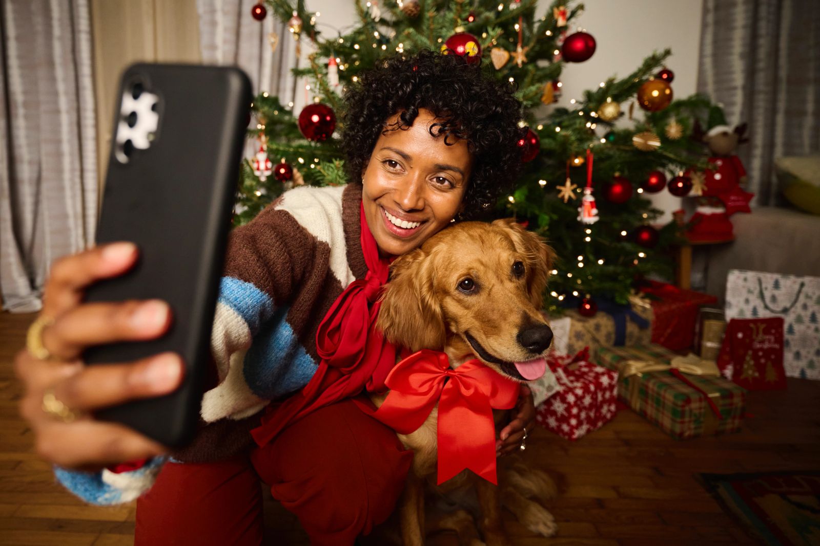 Eine lächelnde Frau macht vor einem geschmückten Weihnachtsbaum ein Selfie mit einem Hund, der eine rote Schleife trägt