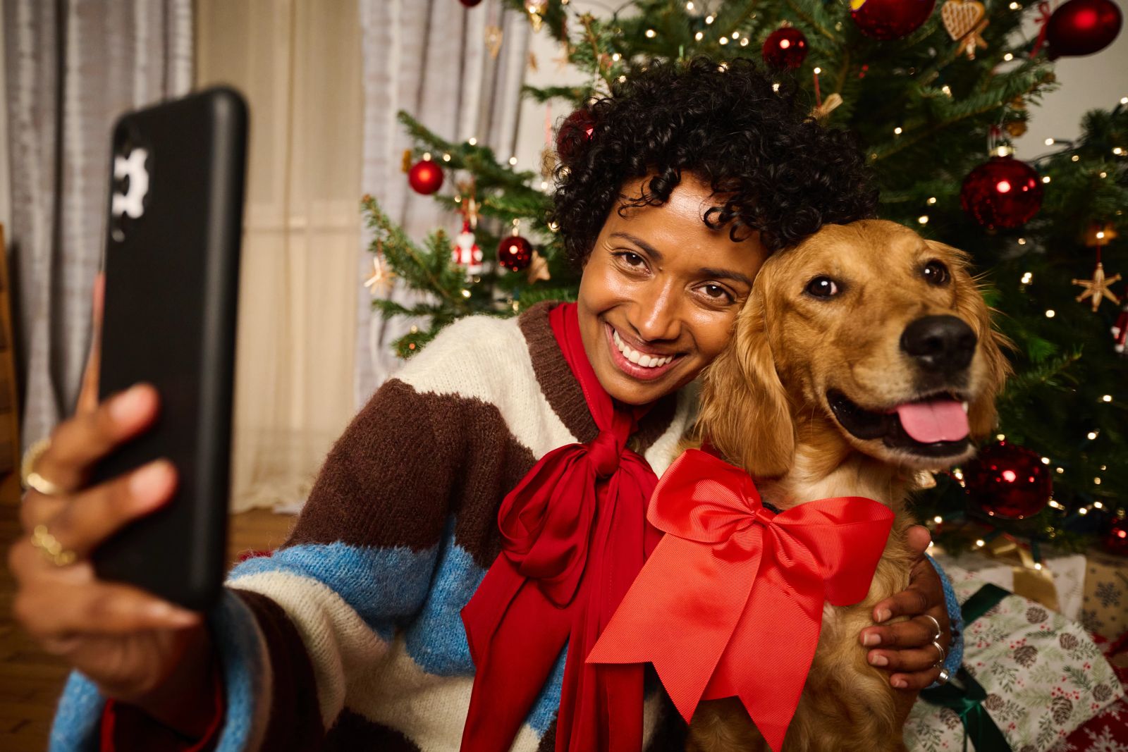 Eine lächelnde Person macht vor einem geschmückten Weihnachtsbaum ein Selfie mit einem Hund, der eine große rote Schleife trägt