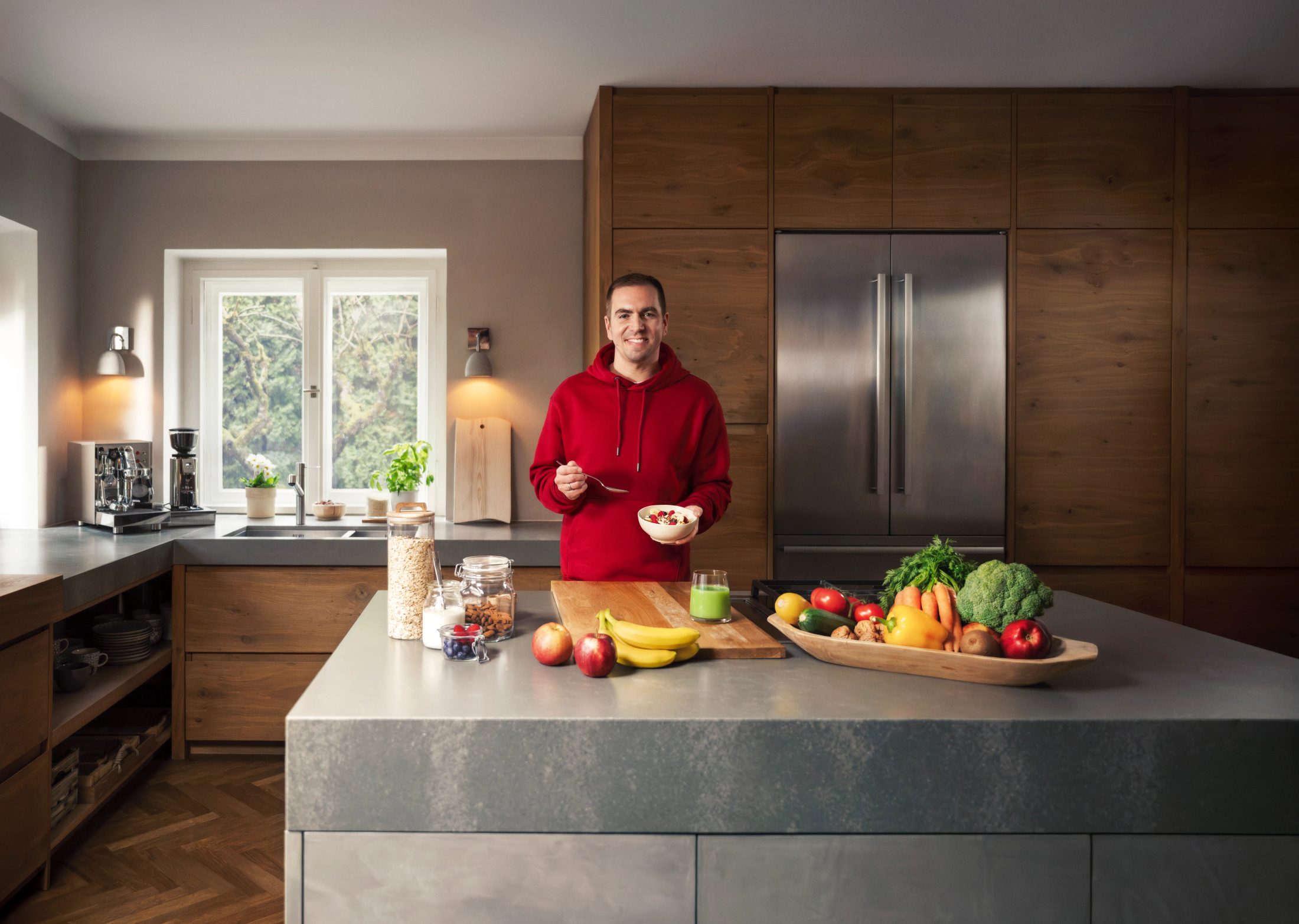 Philipp Lahm steht lächelnd in einer modernen Holzküche und bereitet ein gesundes Müsli zu. Auf der Arbeitsplatte liegen frisches Obst und Gemüse wie Bananen und Brokkoli bereit. Fotografiert von Karl Scholten.