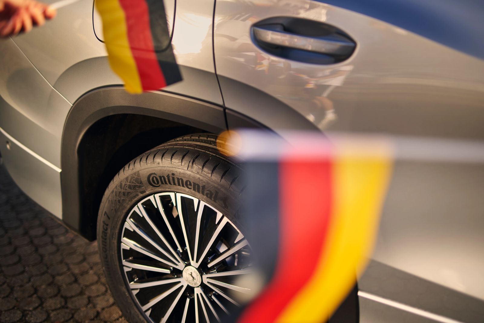 Nahaufnahme eines Autoreifens mit Continental-Schriftzug auf einer Alufelge, im Vordergrund eine unscharfe Deutschlandflagge