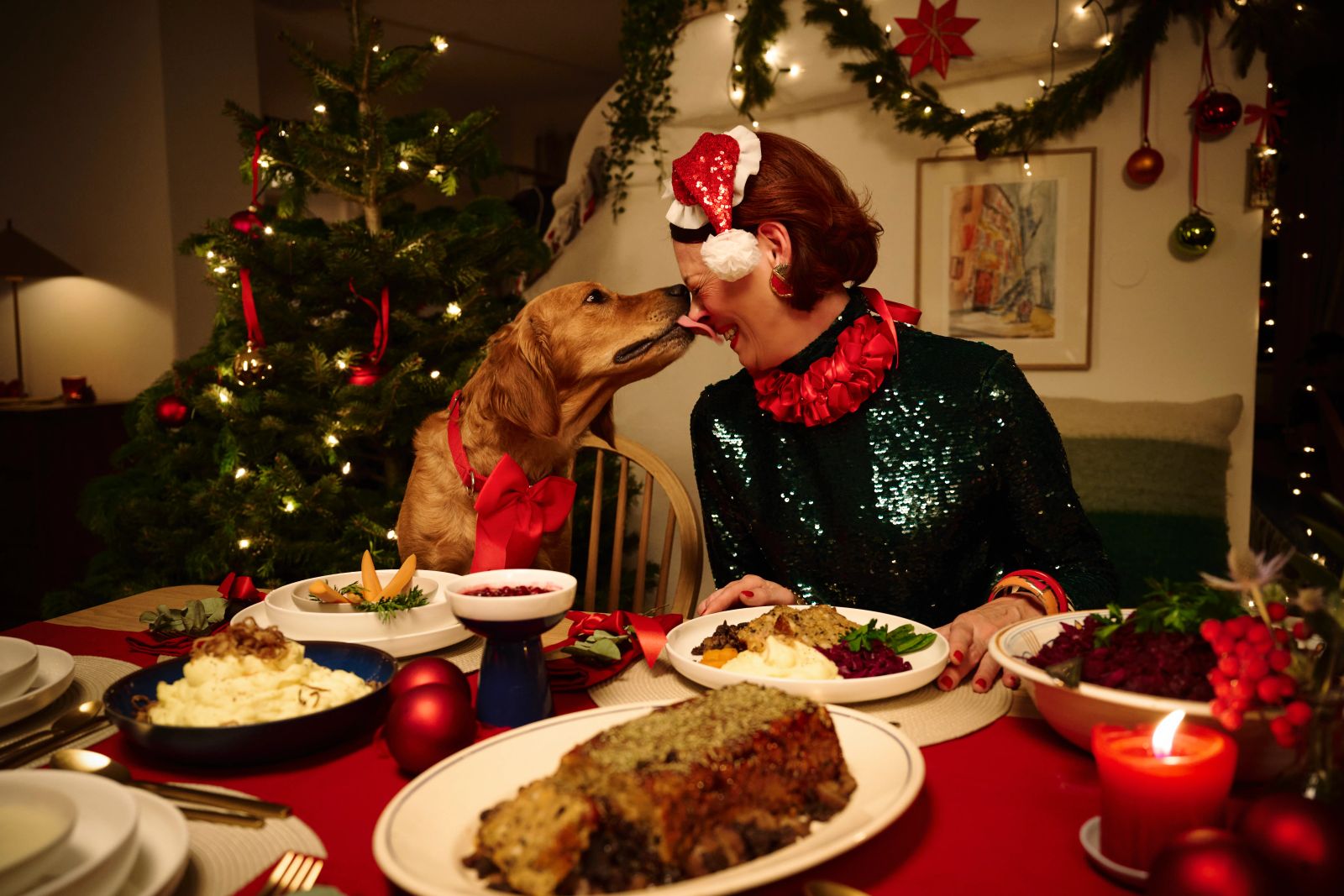 Ein Hund mit einer roten Schleife leckt einer festlich gekleideten Person an einer weihnachtlichen Tafel die Nase
