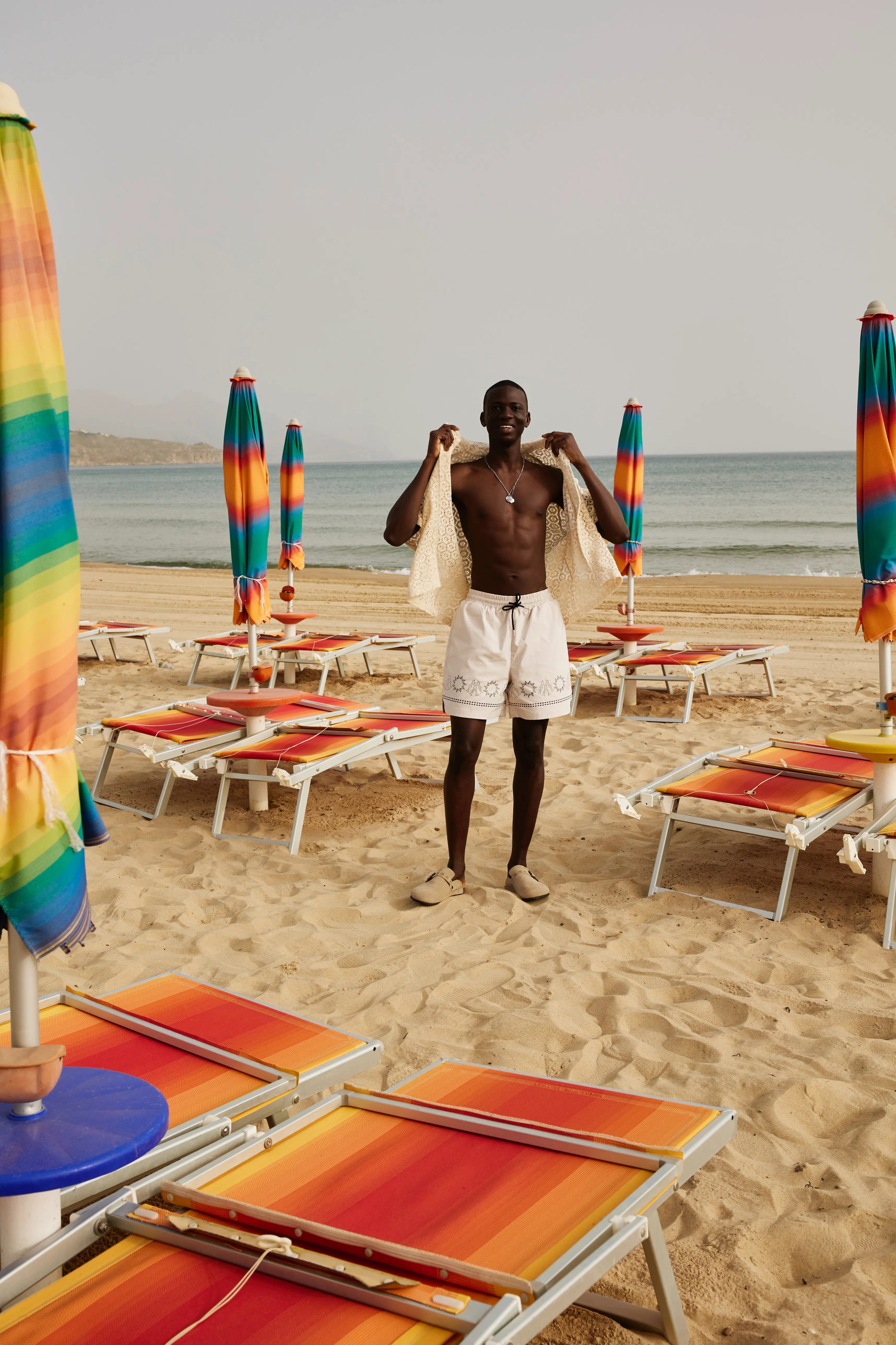 Mann lachend mit gemustertes Mosquets-Oberteil am Strand mit bunten Sonnenschirmen in Sizilien